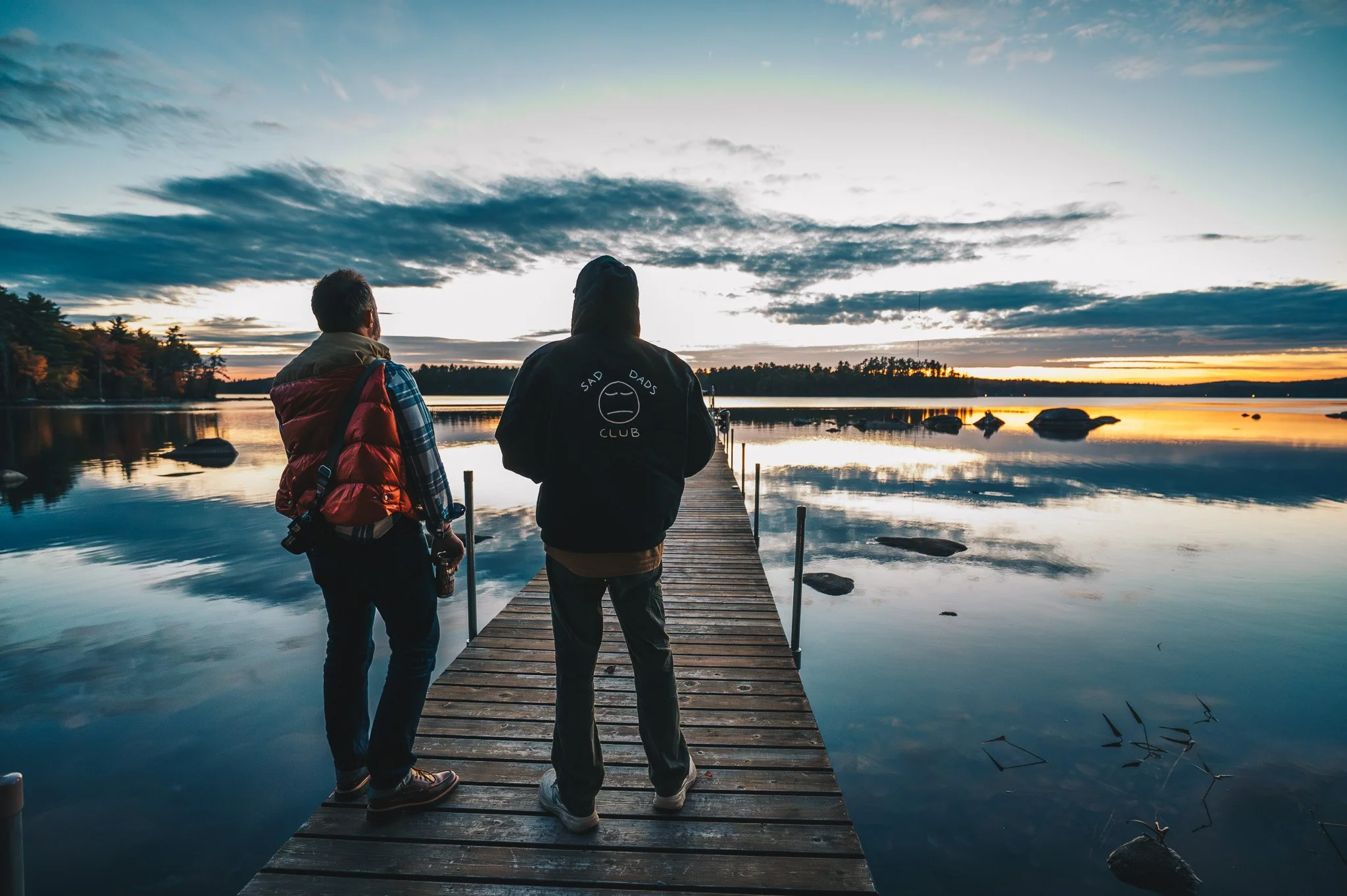 Two men are walking on a wooden dock over a calm body of water at sunset, with trees on the horizon and clouds in the sky.
