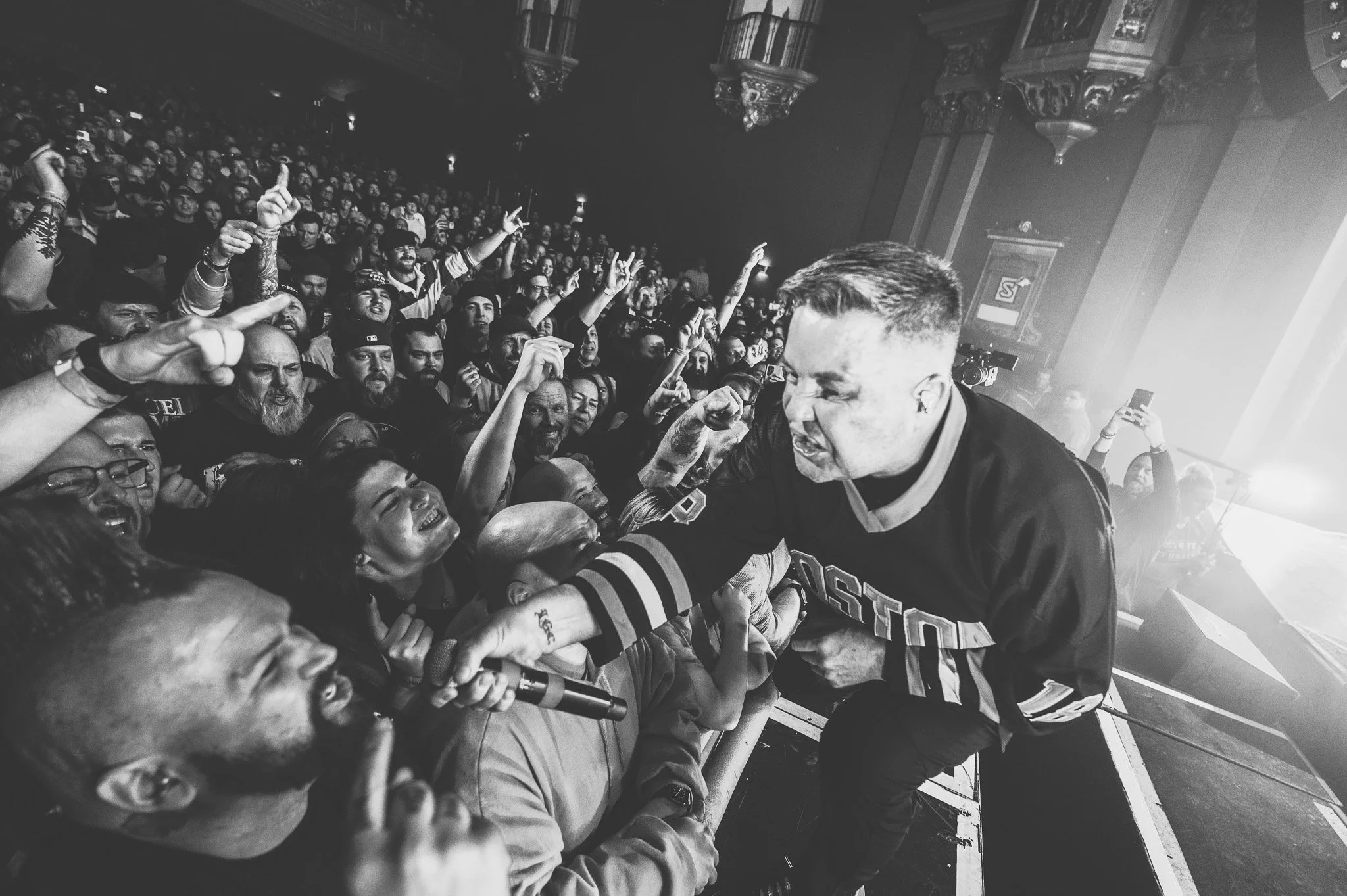 Black and white photo of a crowded concert or event, with a man in the foreground wearing a sports jersey, reaching out and engaging with the audience, who are cheering and raising their hands.