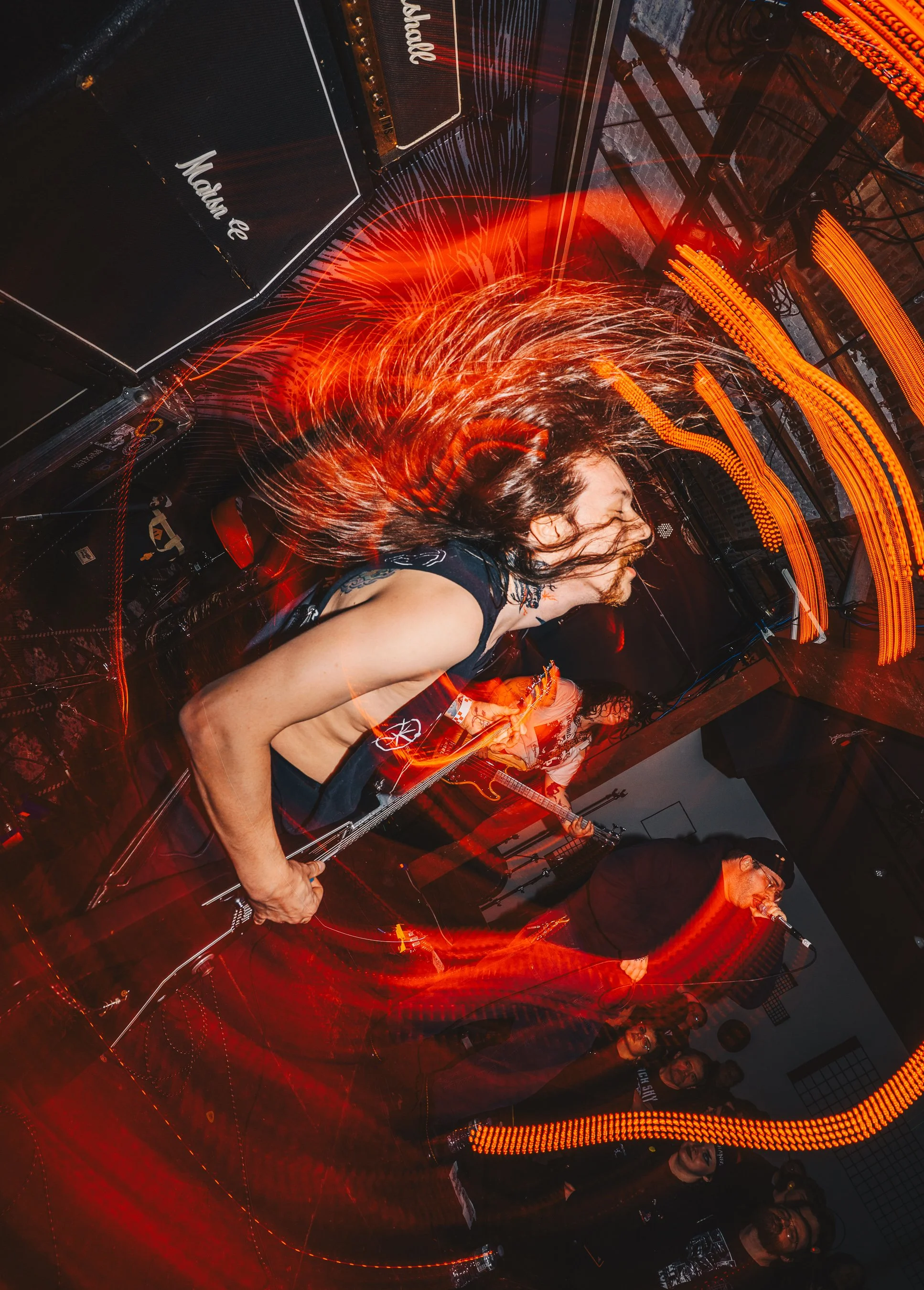 A musician with long hair playing an electric guitar at a live concert, surrounded by red and orange light trails and LED lighting on stage.