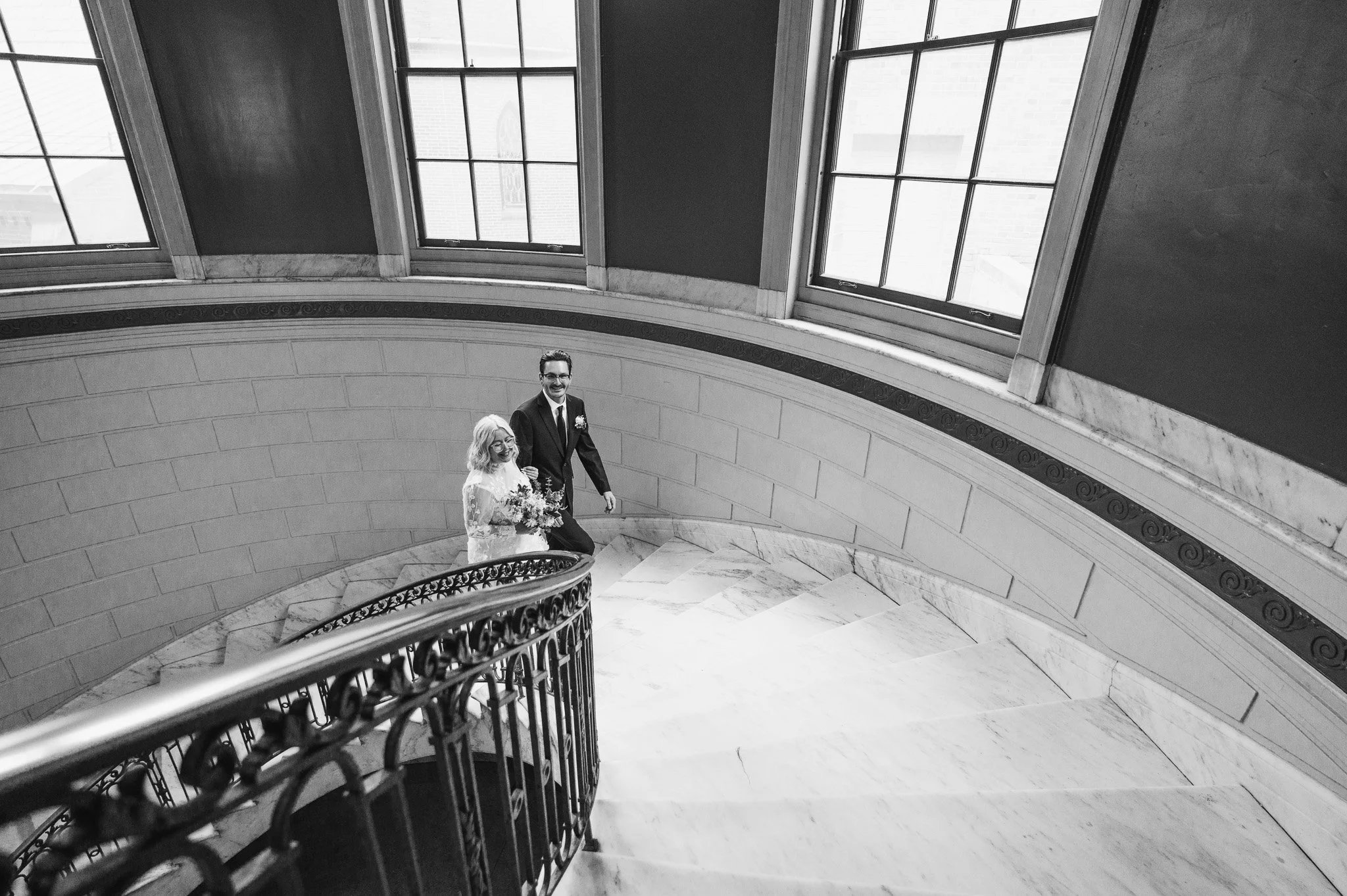 Black and white photograph of a couple dressed in wedding attire standing on a marble staircase inside a building with large windows and decorated railing.