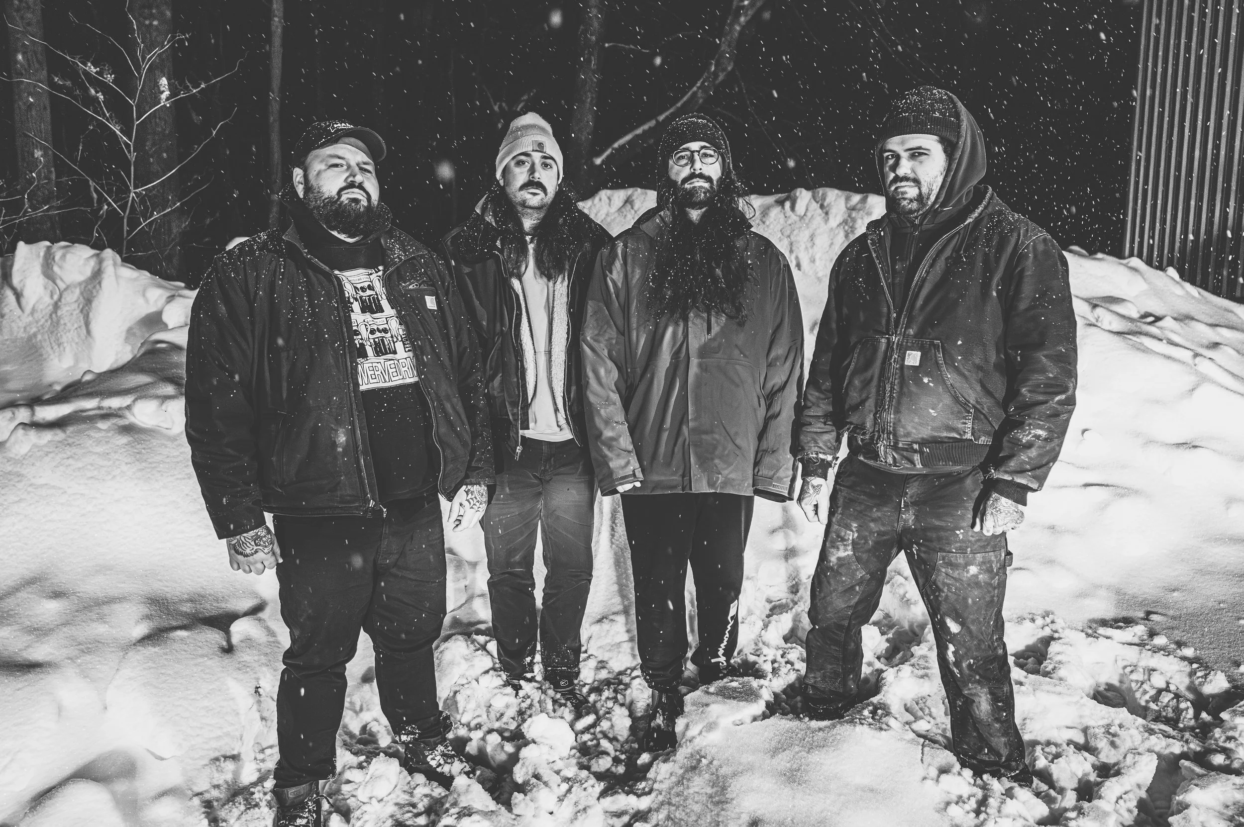 Four men standing in snow with trees and a wooden fence in the background, dressed in winter clothing, with snow falling.