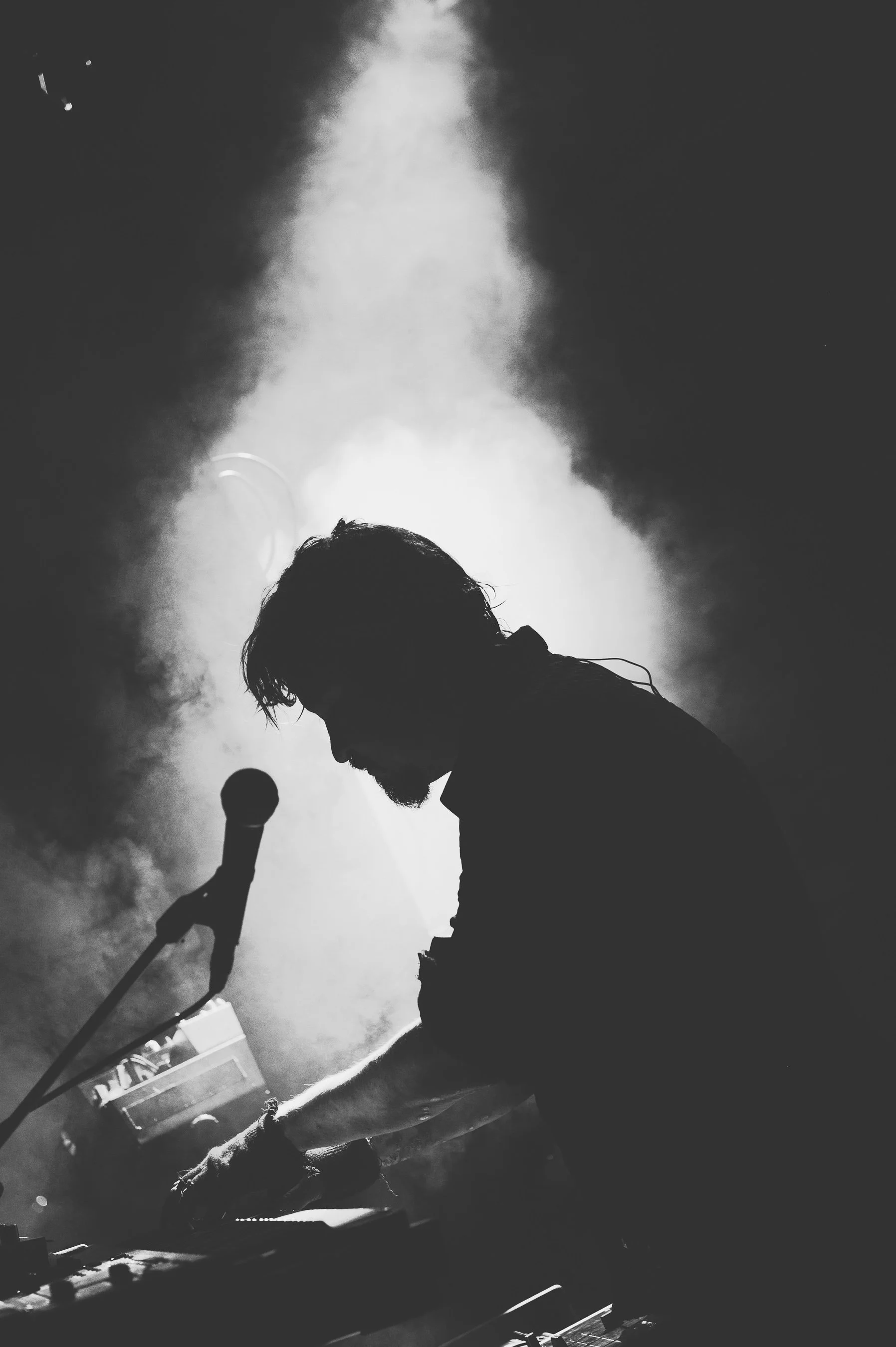 Silhouette of a person with a beard and unkempt hair, holding a microphone, performing on stage with smoke and dramatic lighting.