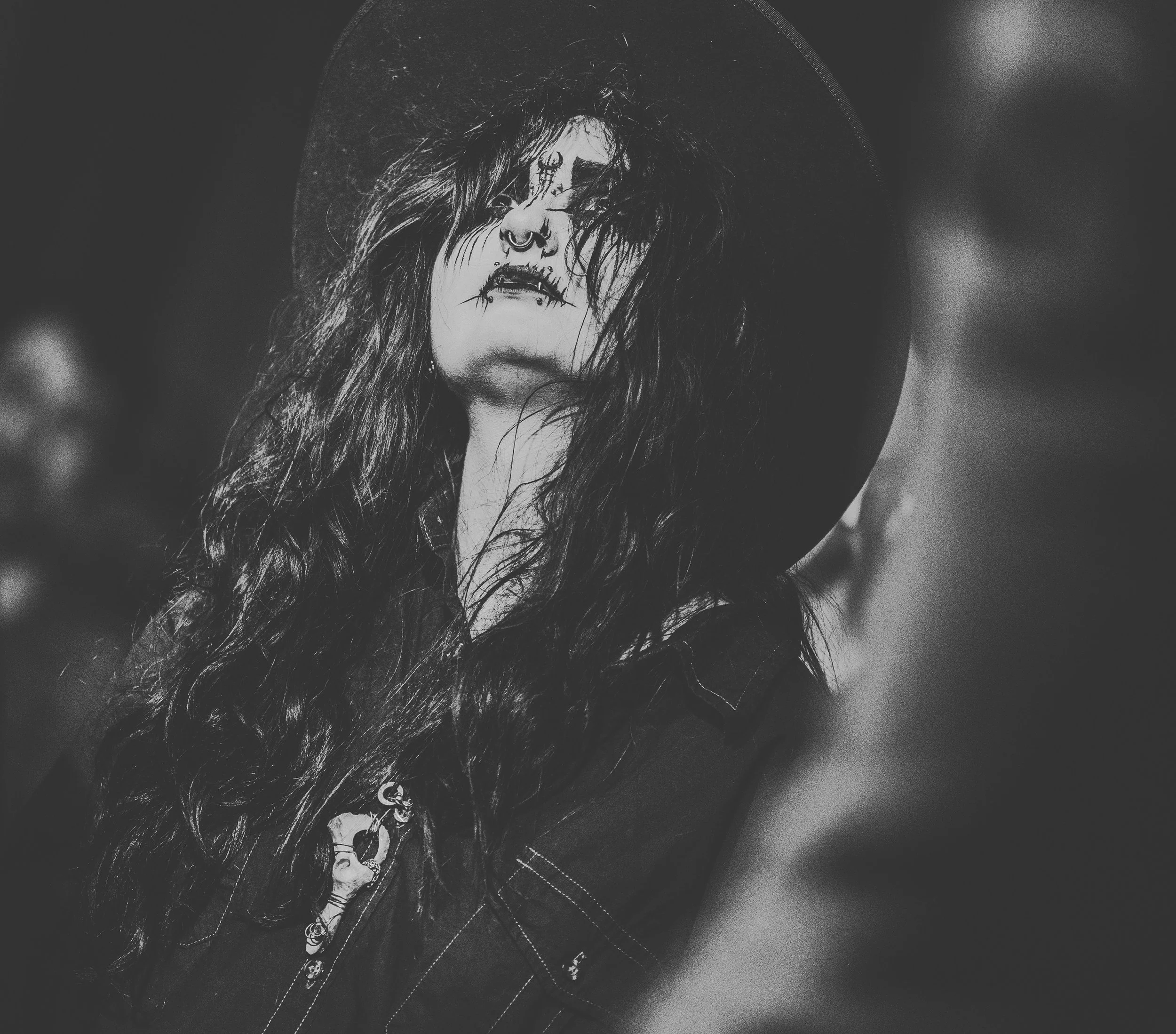 Black and white photograph of a person with long, curly hair, wearing a hat and a bolo tie. The person has facial piercings and is wearing a t-shirt with a graphic design.