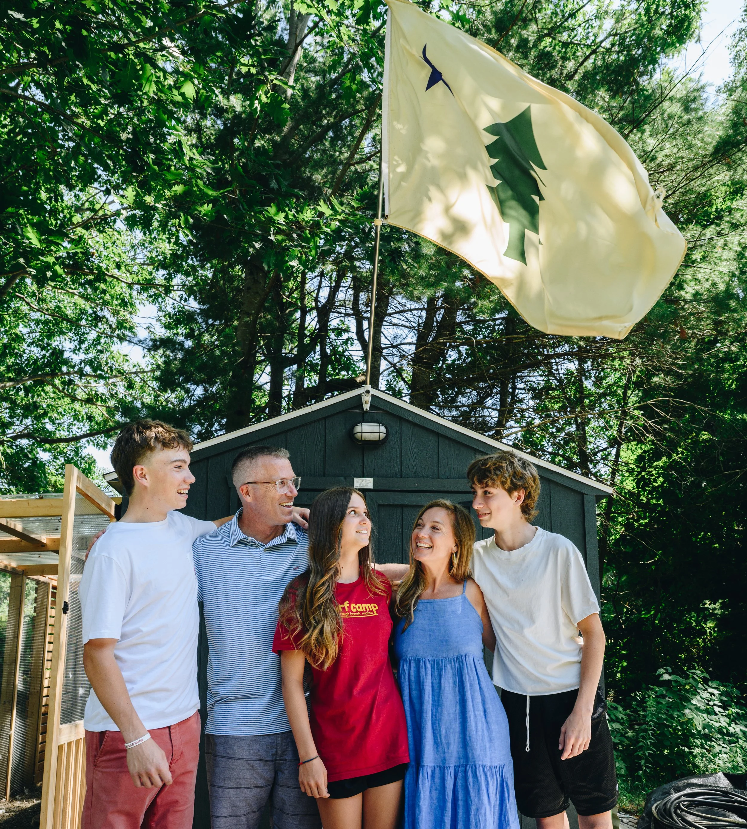 Group of six diverse youth and young adults standing outdoors in front of a black shed, smiling and talking, with a yellow flag featuring a green pine tree flying above them, lush green trees overhead.
