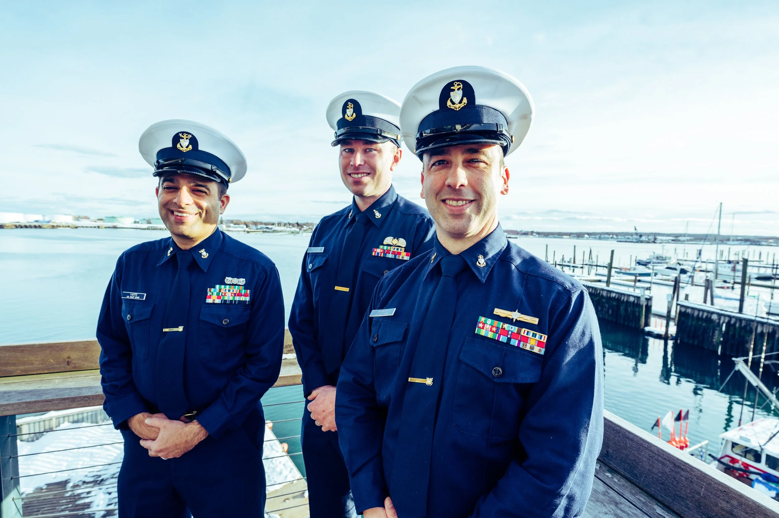 Three men in navy uniforms standing outdoors near water, smiling at the camera, with a marina and boats in the background.
