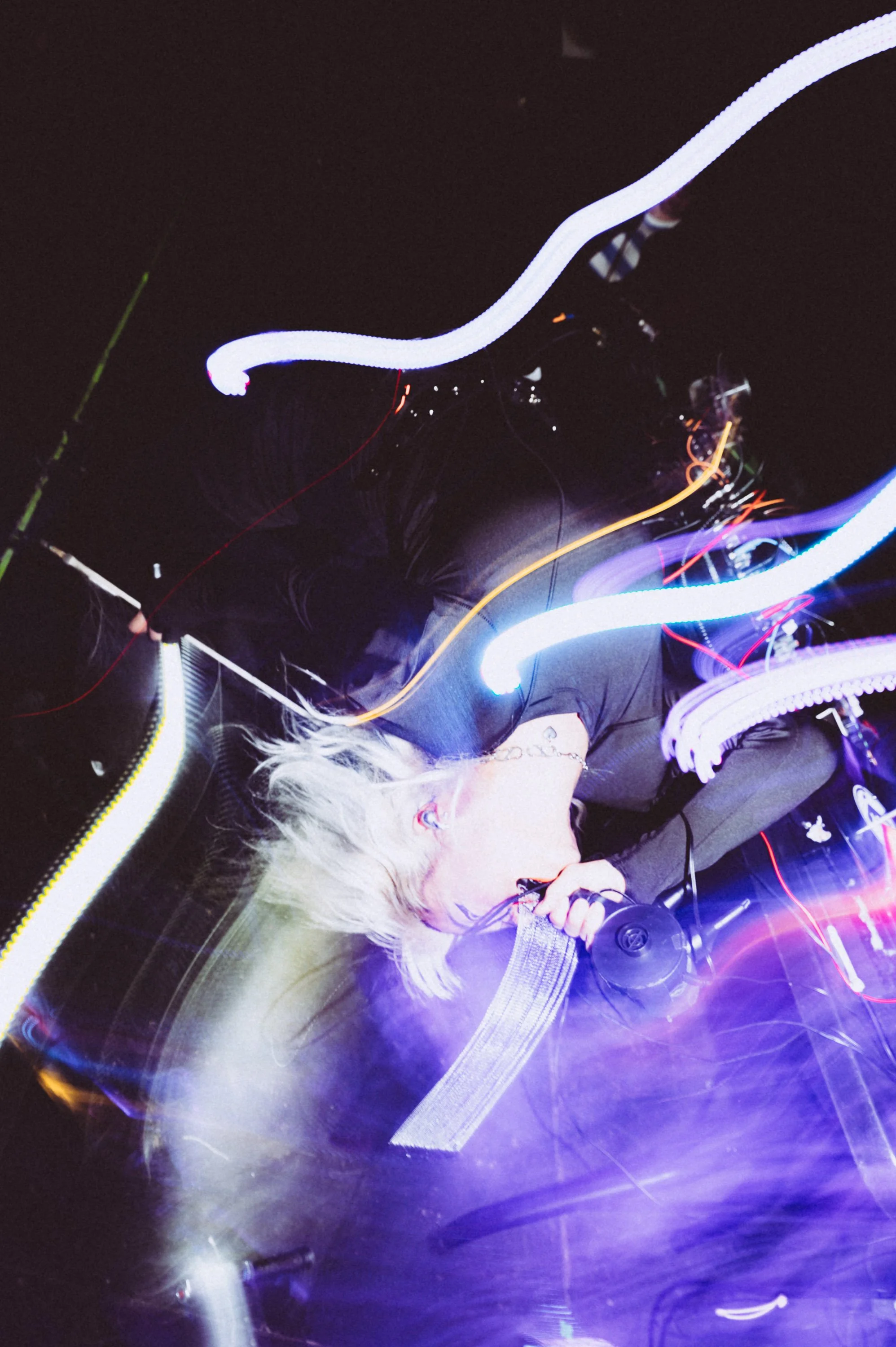 A person with glasses and platinum blonde hair blowing into a microphone, with colorful light trails and vibrant neon lighting creating a dynamic, energetic atmosphere.