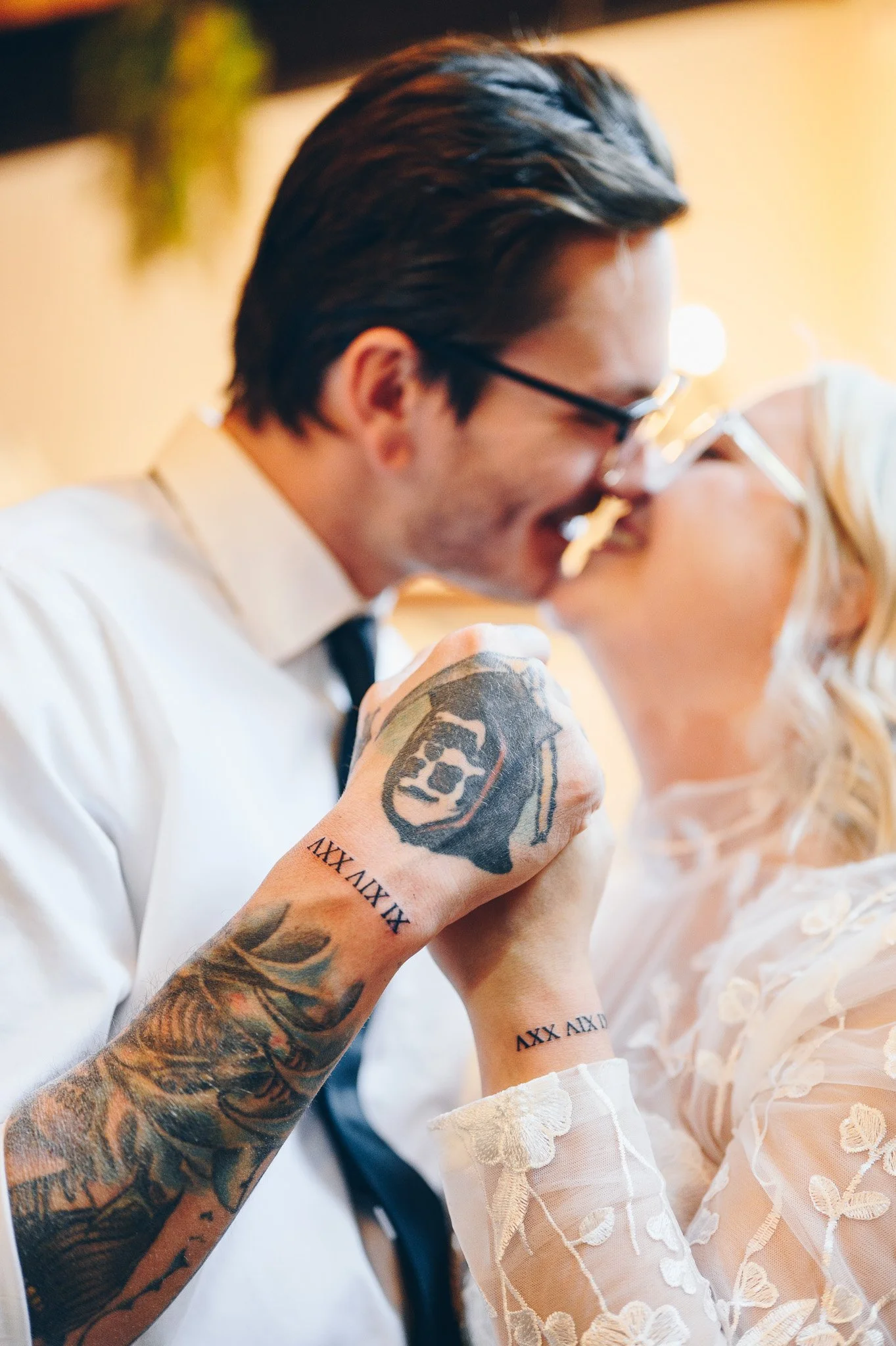 A man with dark slicked-back hair and glasses smiling, with tattooed arms, wearing a white shirt and tie, holding his arm up in front of a smiling woman with blonde hair wearing a light-colored dress, both appearing to be at a special event.