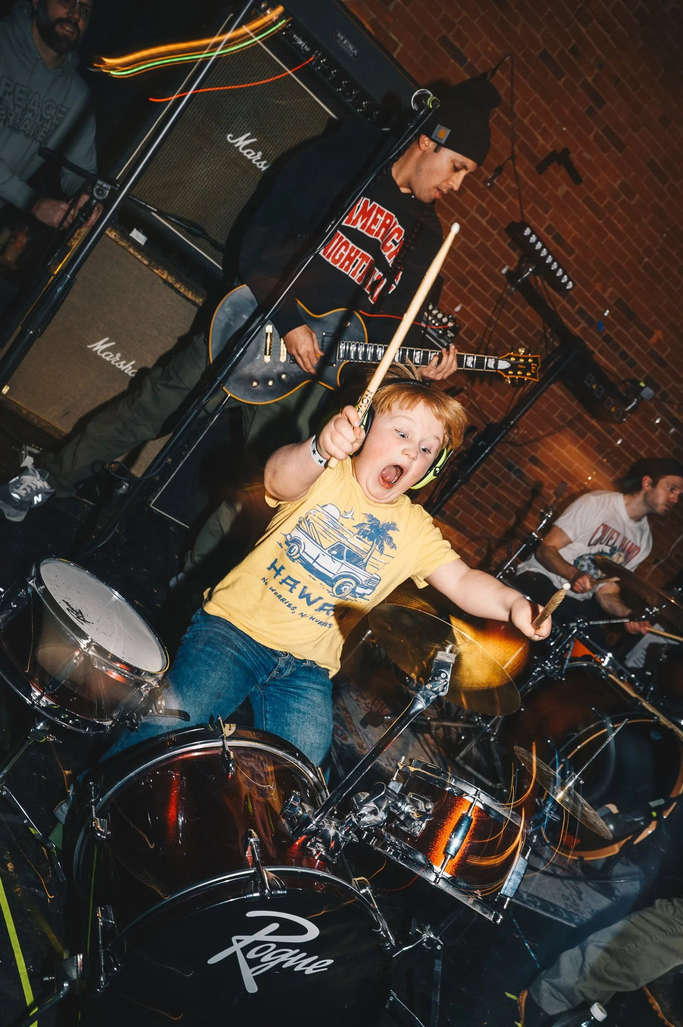 A young boy with red hair wearing a yellow t-shirt and blue jeans is playing drums, with an expression of excitement. Behind him, a musician in a black beanie and hoodie is playing an electric guitar, with a brick wall and stage lighting in the backg