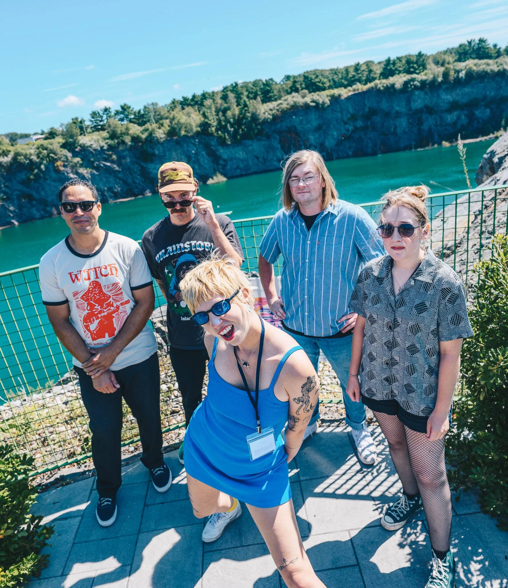 Group of five young adults outdoors by a river with a cliff and green trees in the background, posing for a photo on a sunny day.