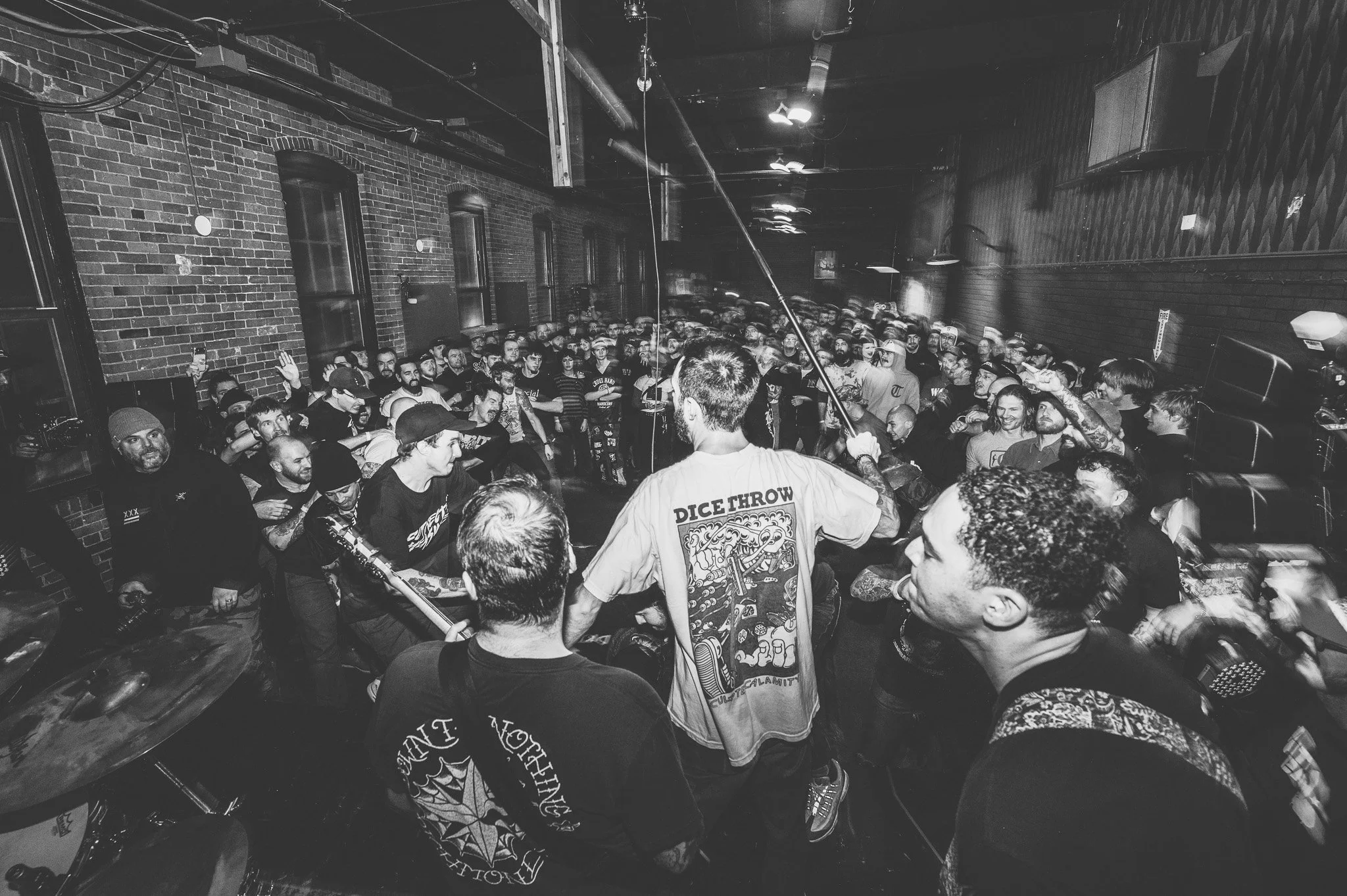 A large crowd of people gathered in a dimly lit indoor space with exposed brick walls and industrial lighting, attending a music event or concert.
