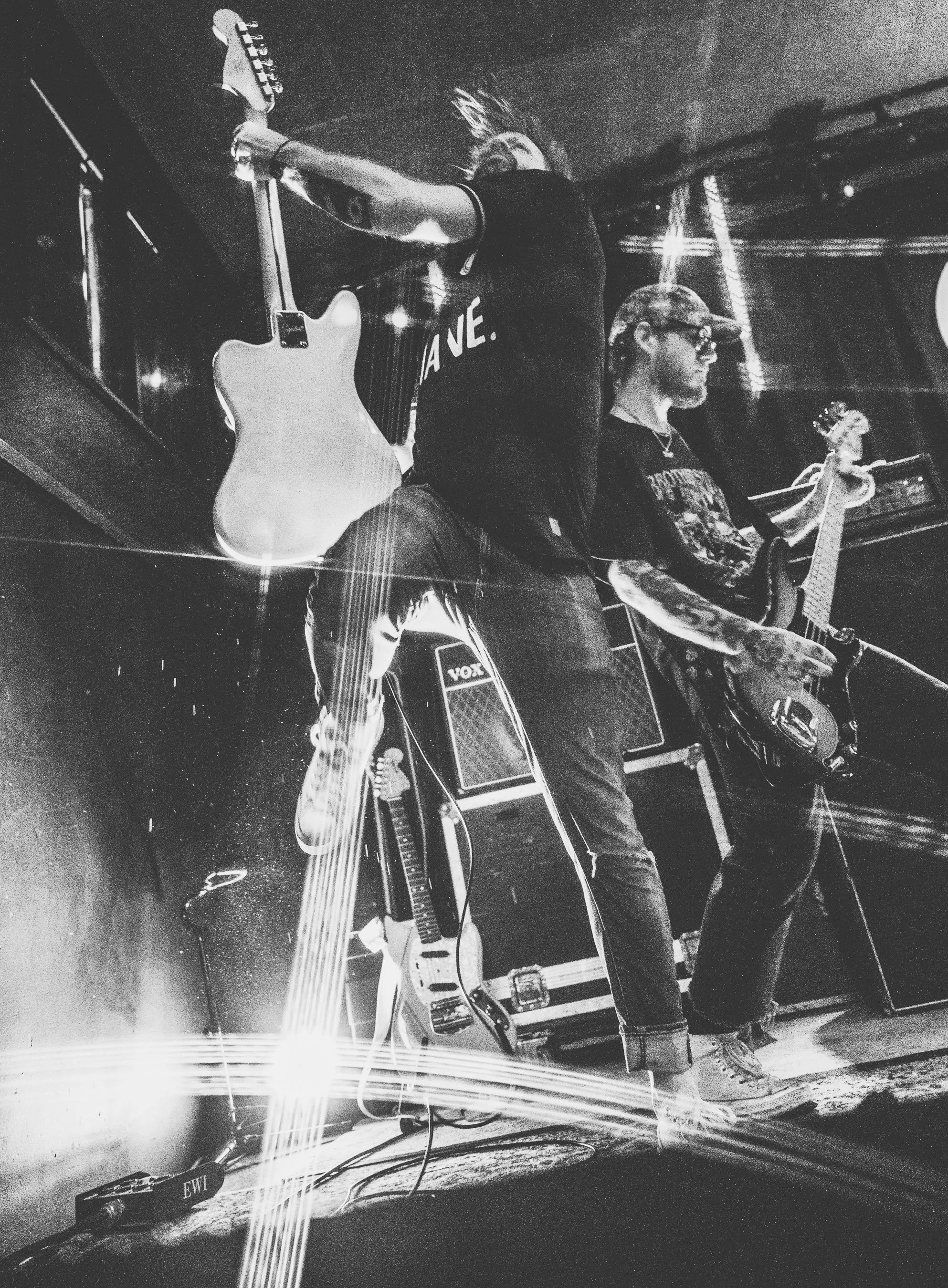 Two musicians performing with guitars in a dimly lit setting, one with a hat and glasses, the other with spiked hair, surrounded by guitar amplifiers and equipment.