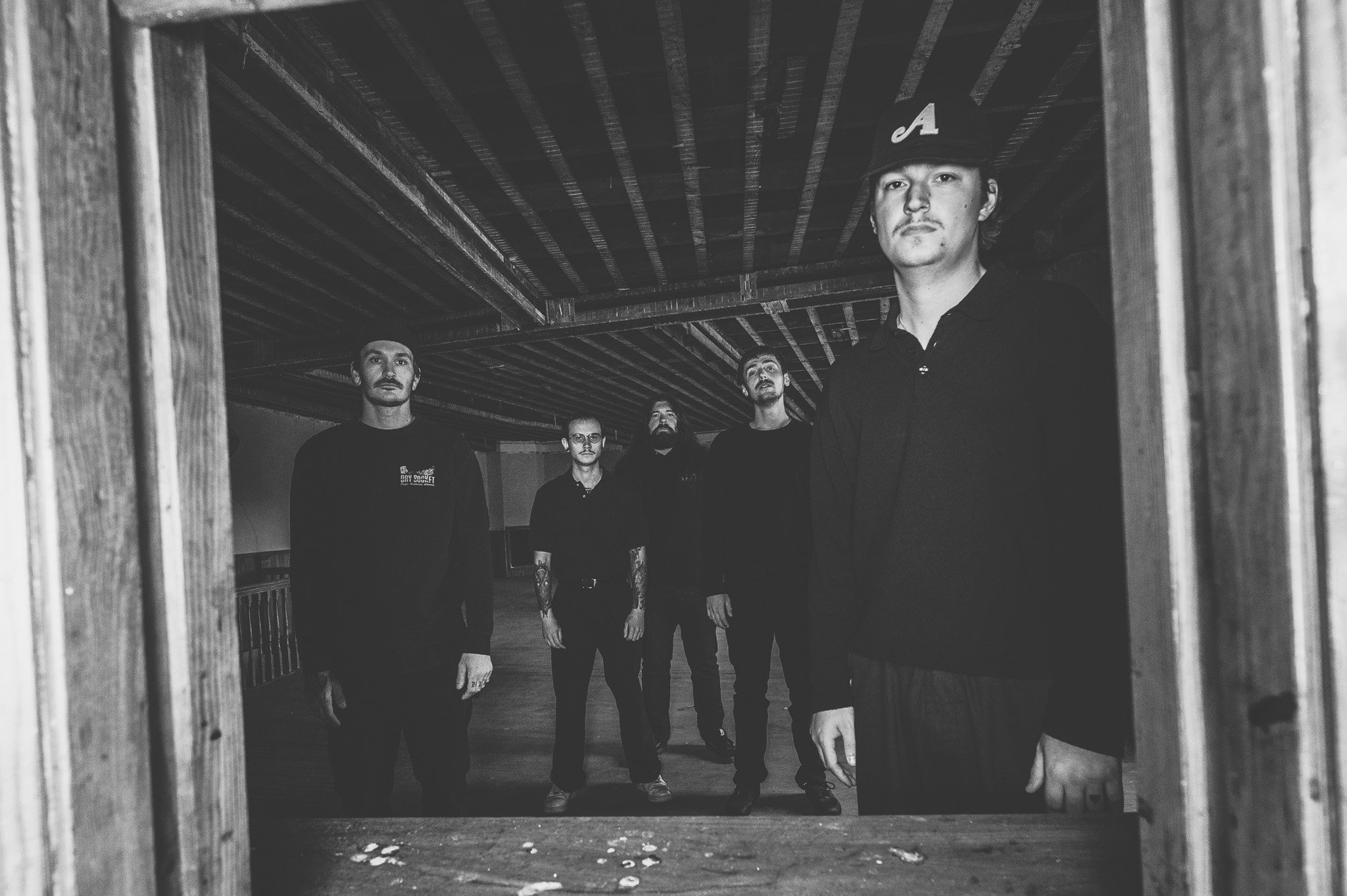 A group of five men standing in a dimly lit wooden structure, looking towards the camera. One man in the foreground is wearing a cap with the letter 'A' on it.