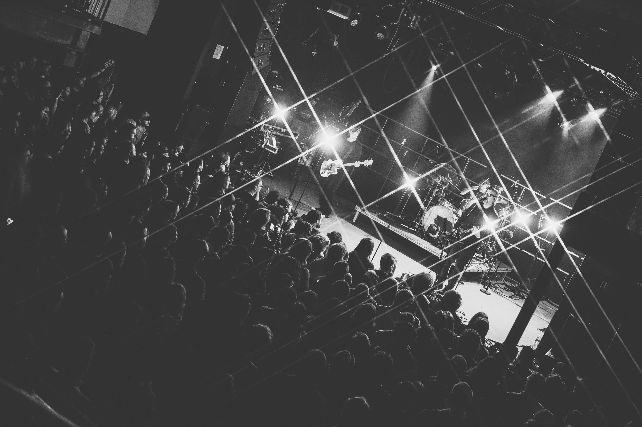 Black and white photo of a live music concert with an audience facing the stage. The stage is illuminated with bright lights and lasers, with visible musical instruments, including a guitarist and drummer.