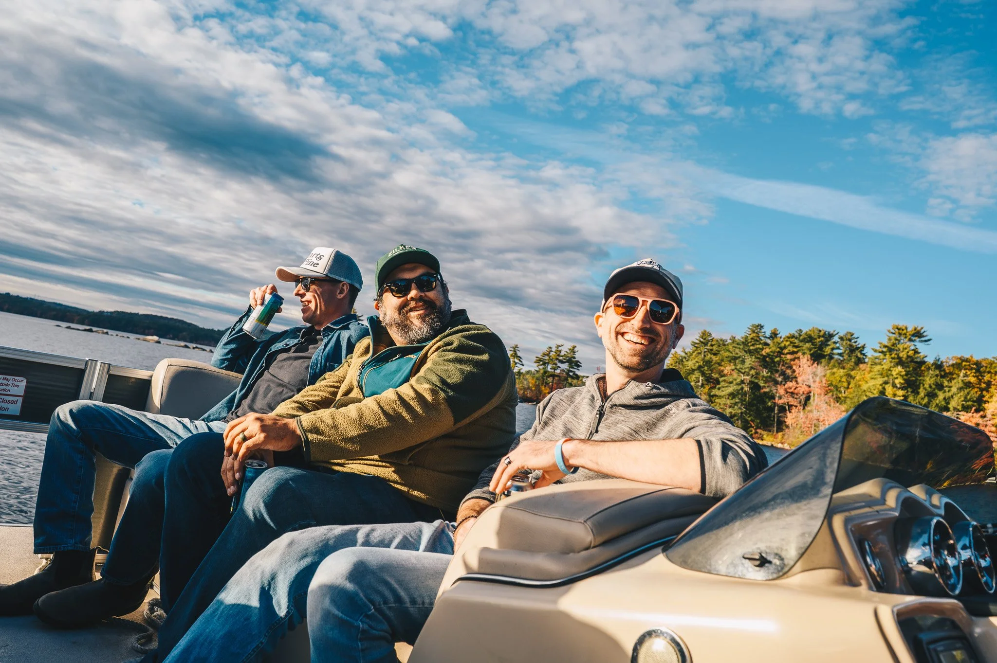 Three men riding on a boat on a lake during daytime with blue sky and scattered clouds, trees with fall foliage in the background, smiling and enjoying the ride.