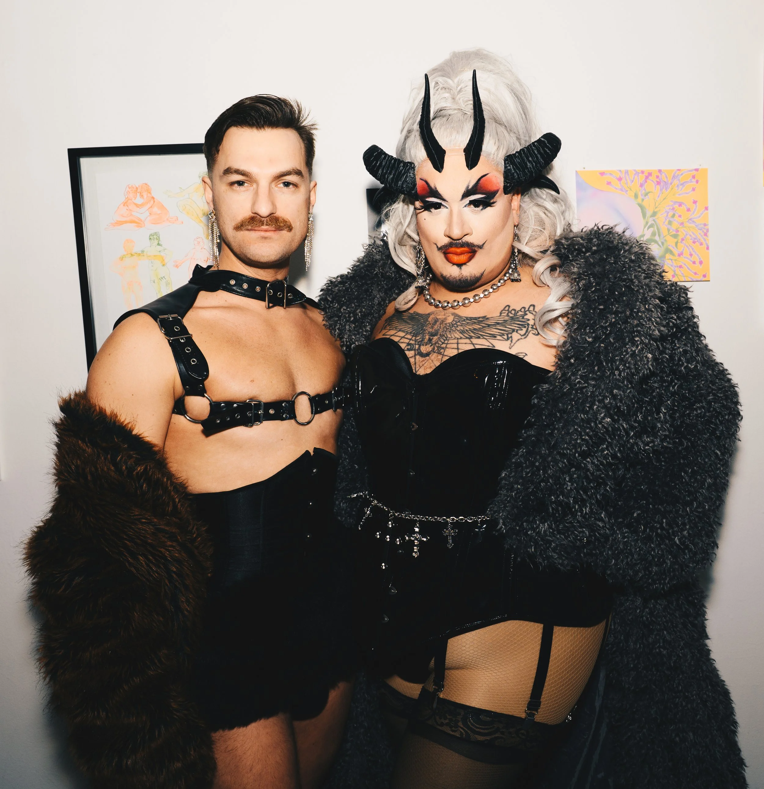 Two individuals dressed in bold, revealing outfits with heavy makeup and accessories, standing against a plain wall with framed artwork in the background.