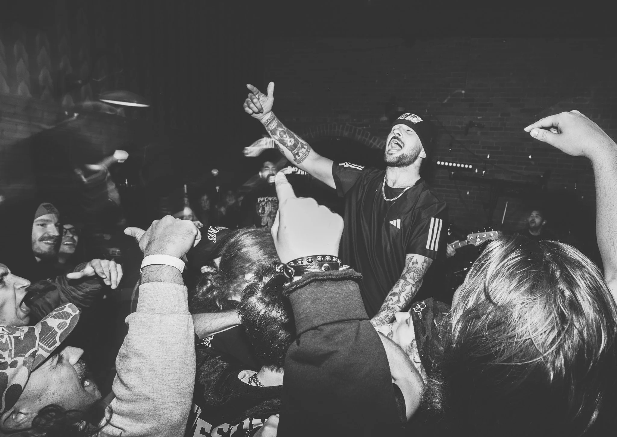 A lively indoor gathering of people celebrating with a man in the center raising his arms, wearing a beanie, tattoos, and a sports jersey, surrounded by smiling friends, some in hooded sweatshirts, with a brick wall background.