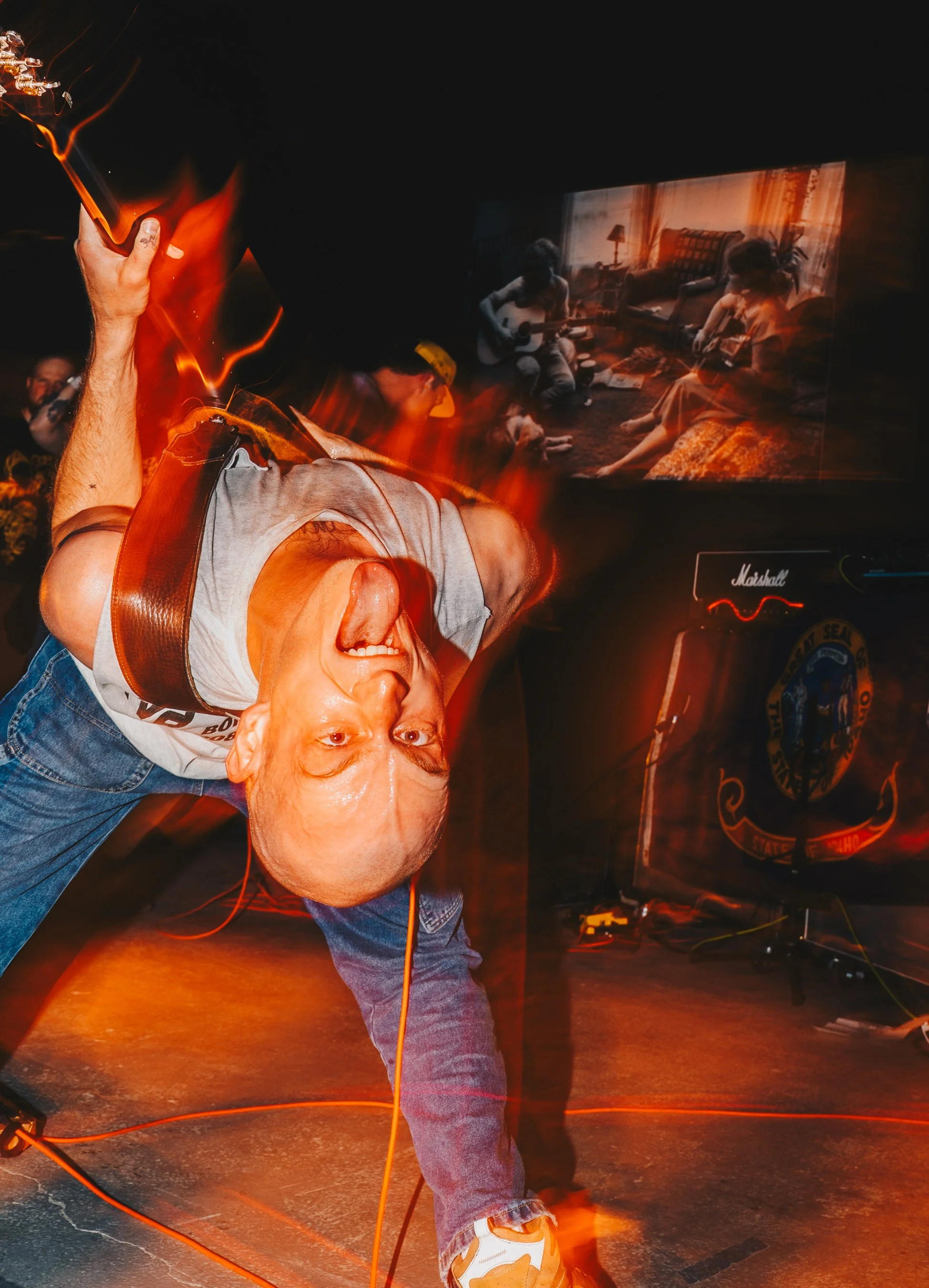A man with a shaved head and a jean jacket leaning forward with his mouth open, dancing in front of a dark background with a screen displaying people playing guitars and a Marshall amplifier visible. The image has a warm, fiery glow.