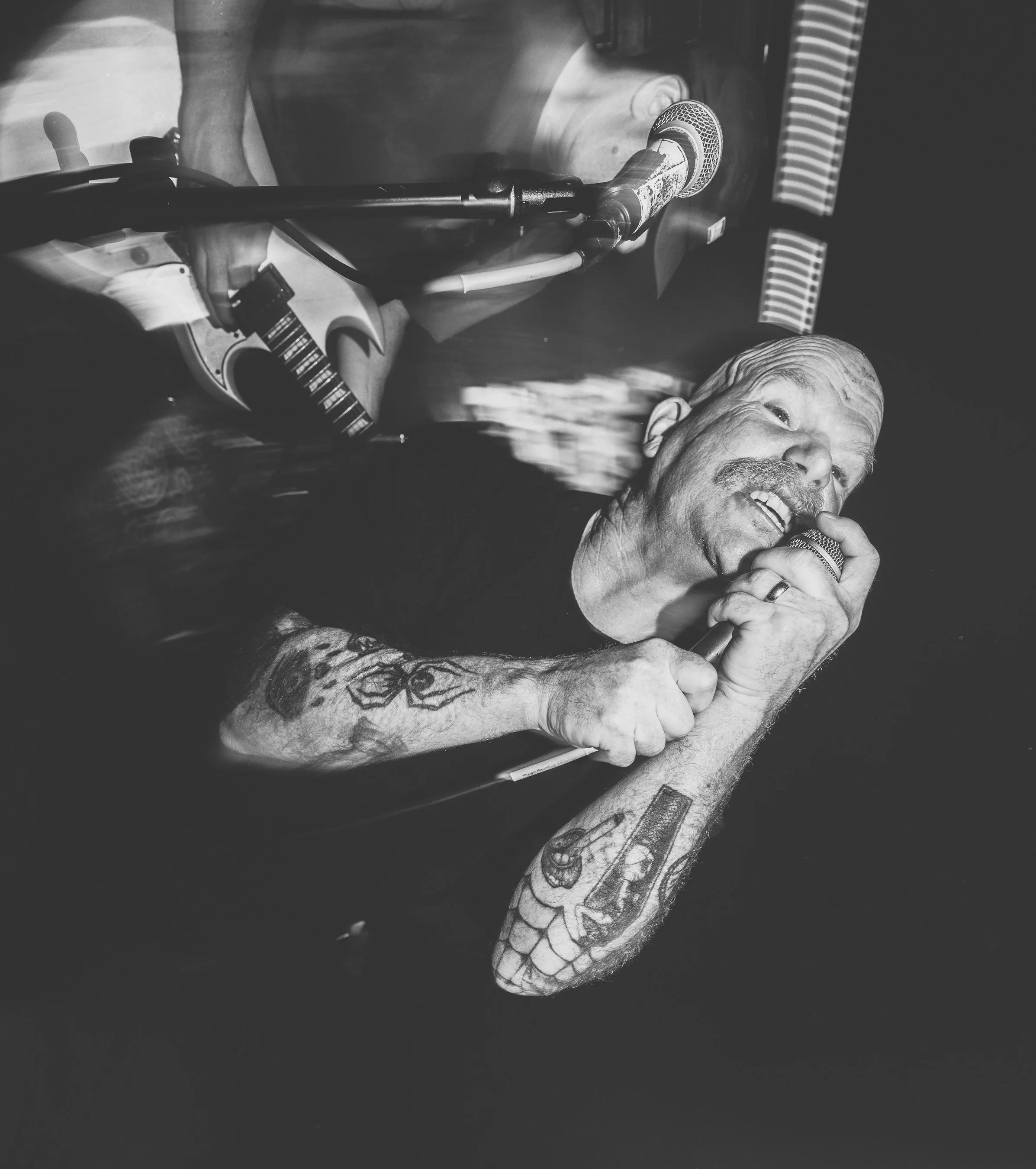 An elderly man with tattoos on his arm is smiling while holding a microphone near his mouth, possibly singing or speaking, with the background showing speakers and equipment.