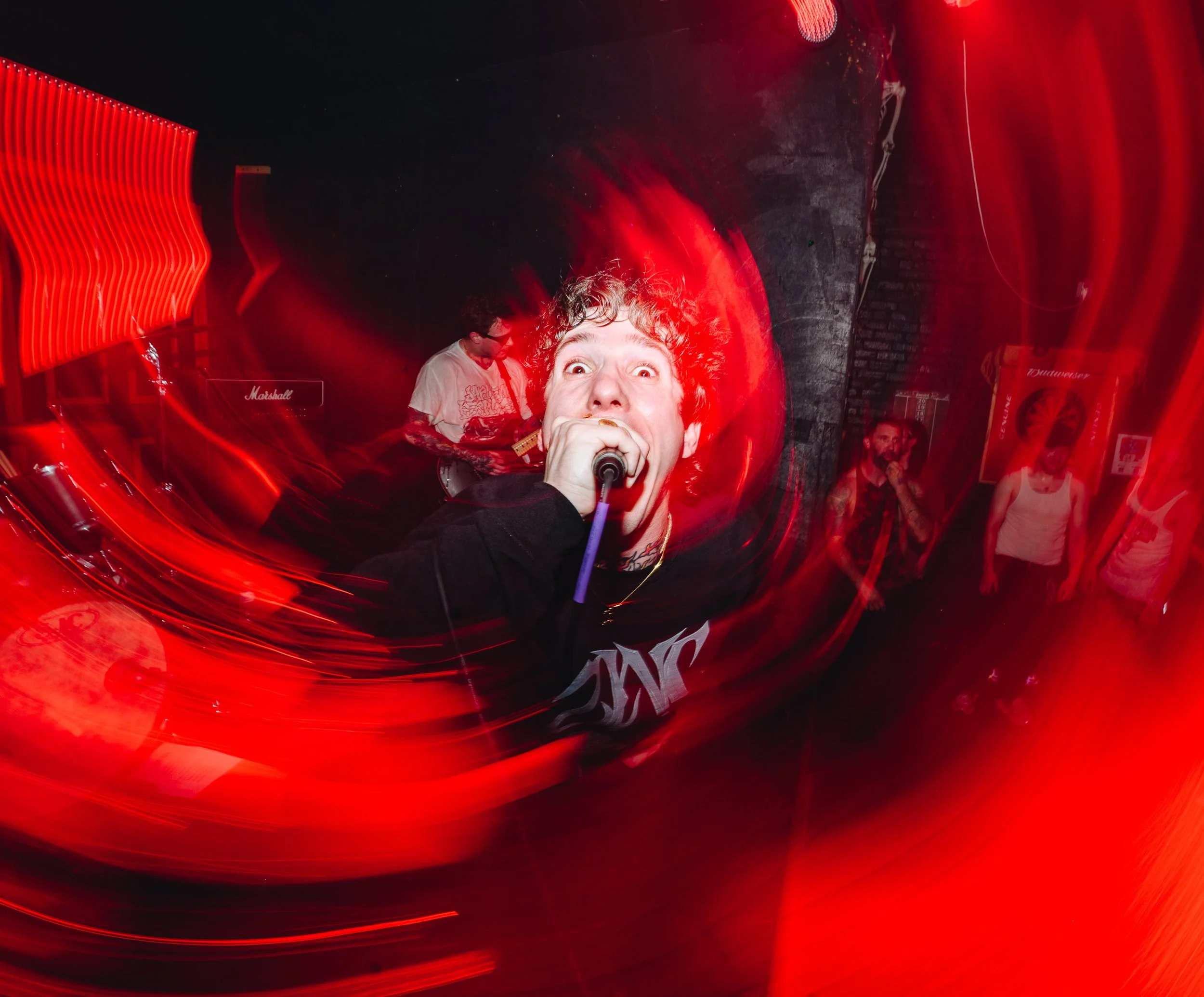A person with curly hair singing into a microphone in a dimly lit setting with red light streaks around them. There are other people visible in the background.