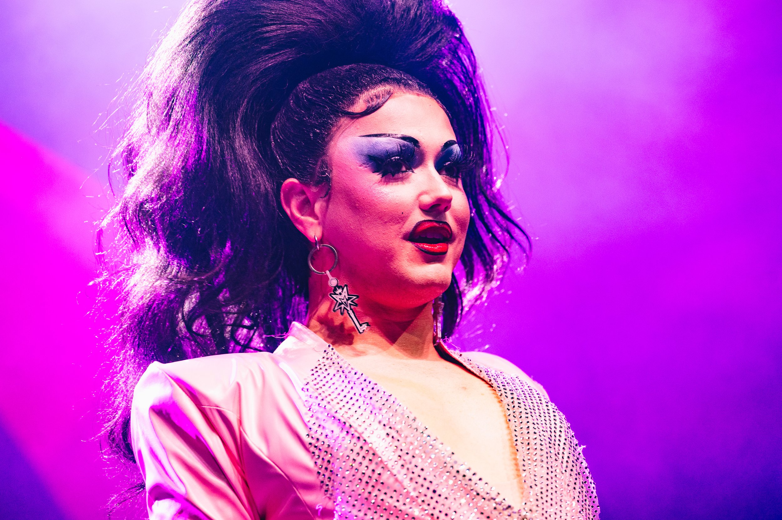 A woman with dark hair styled in an exaggerated, voluminous updo, wearing bold makeup with dark eyeshadow and lipstick, and bright earrings, against a purple background.