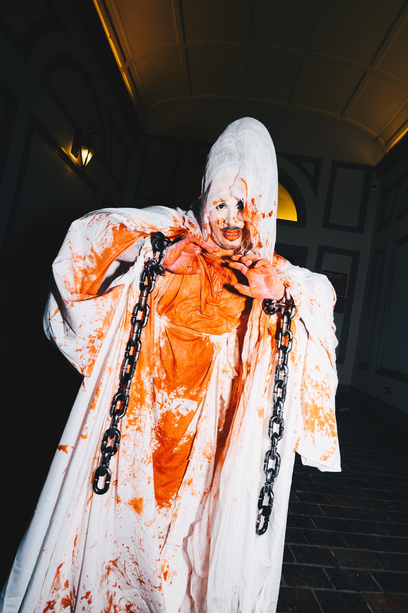 Person dressed as an angel with bloodstains on white robe and chains, standing indoors with dark walls and ceiling, facing the camera.