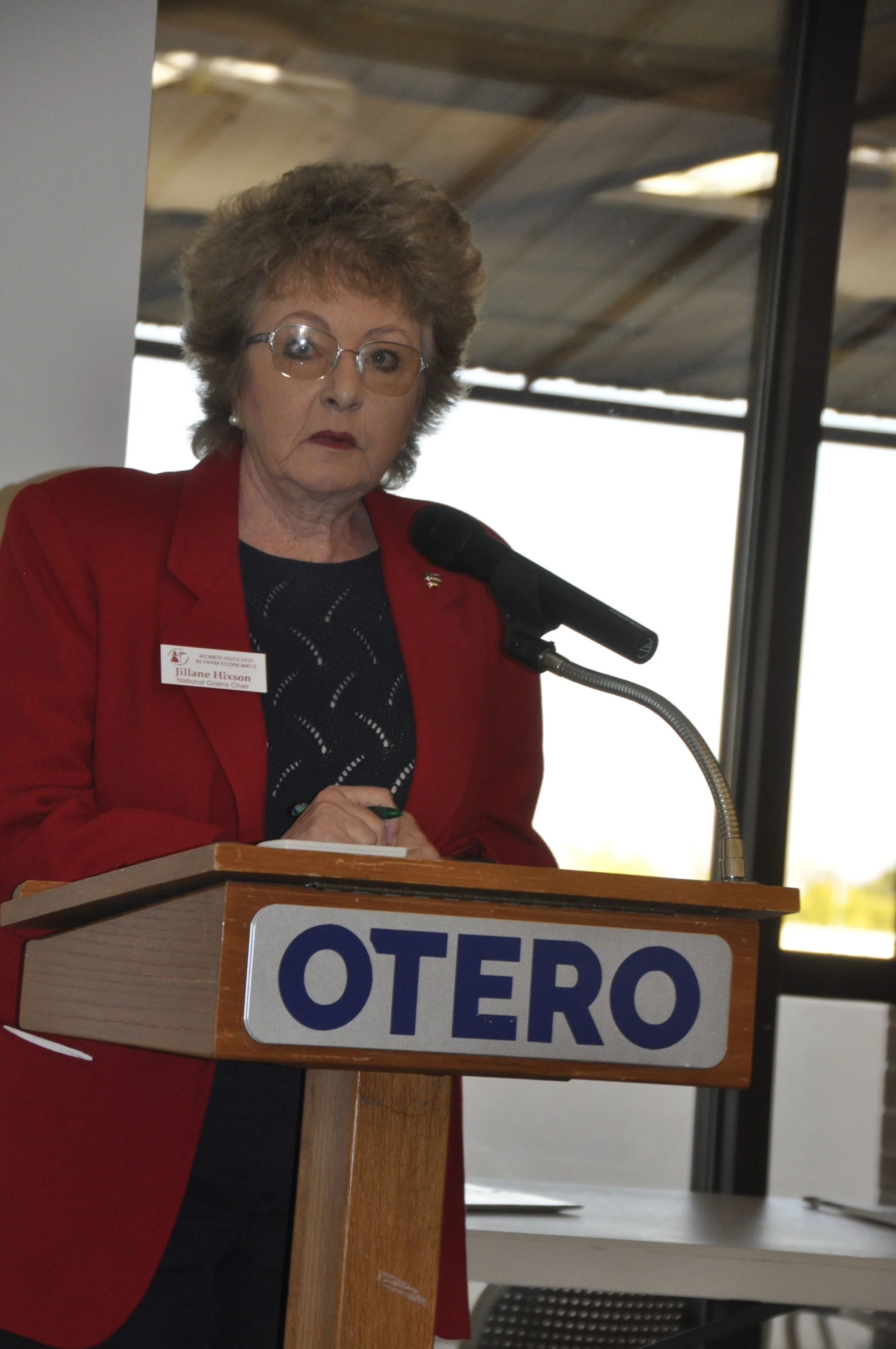 A woman with short, curly hair and glasses wearing a red blazer and a black shirt, standing at a wooden podium labeled 'OTERO', speaking into a microphone.