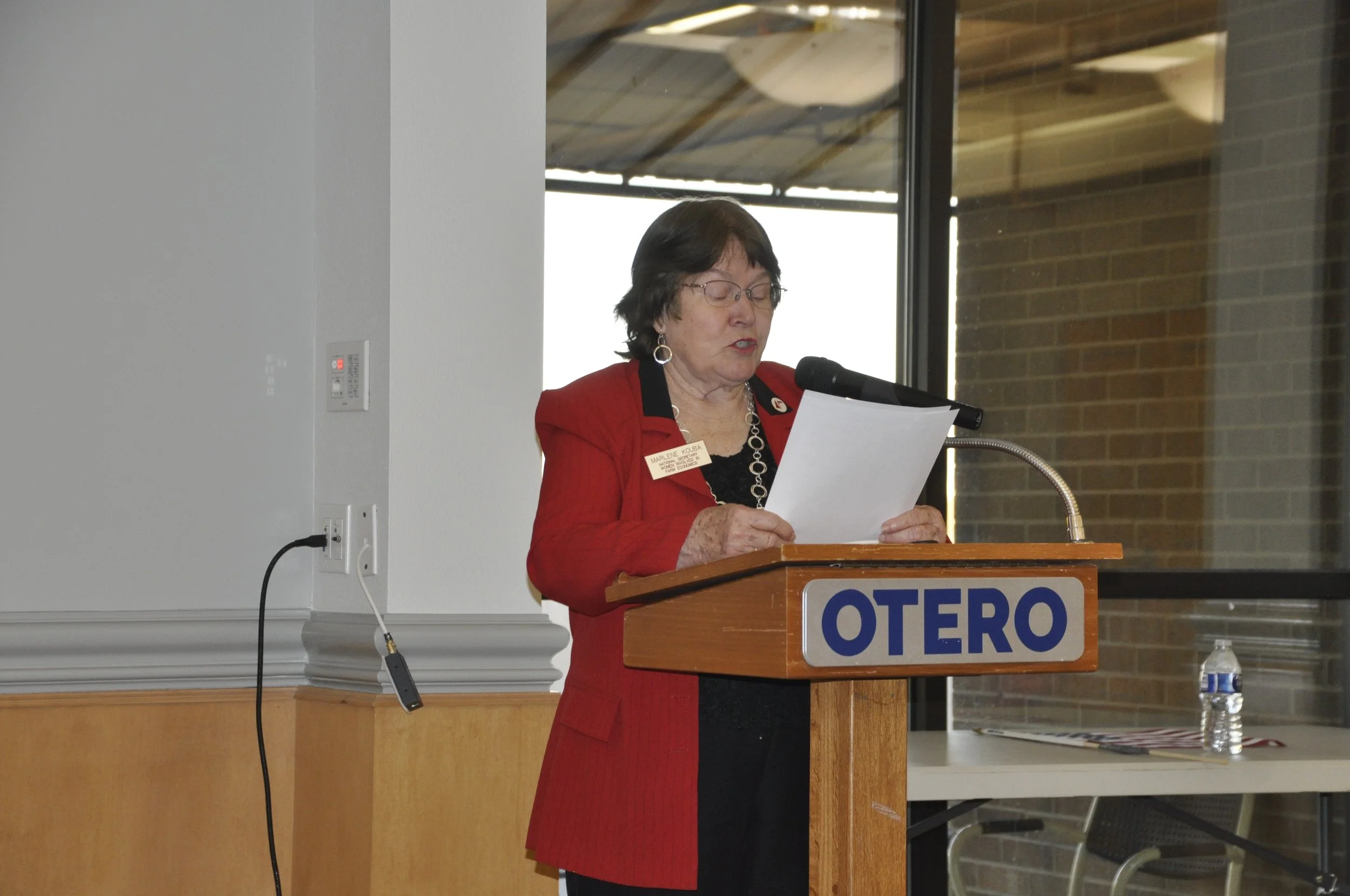 A woman wearing glasses and a red jacket, standing at a podium labeled "OTERO," reading from a piece of paper during an indoor event.