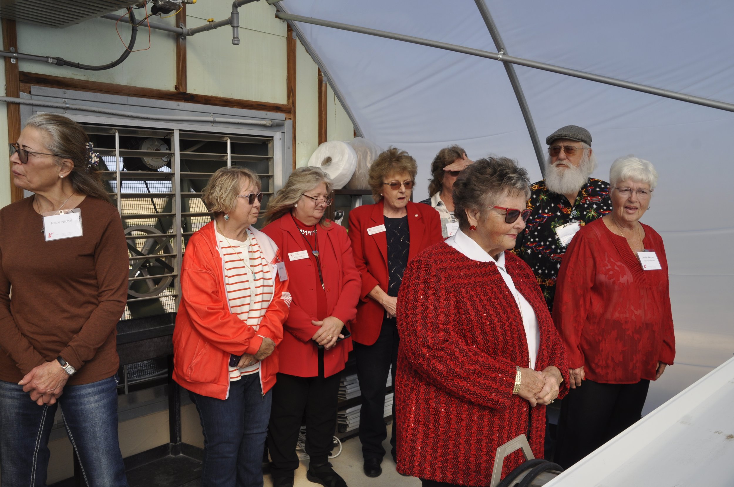 A group of seniors, mostly women, dressed in red and festive attire, are standing inside a greenhouse, looking at and listening to a person or display not visible in the photo.