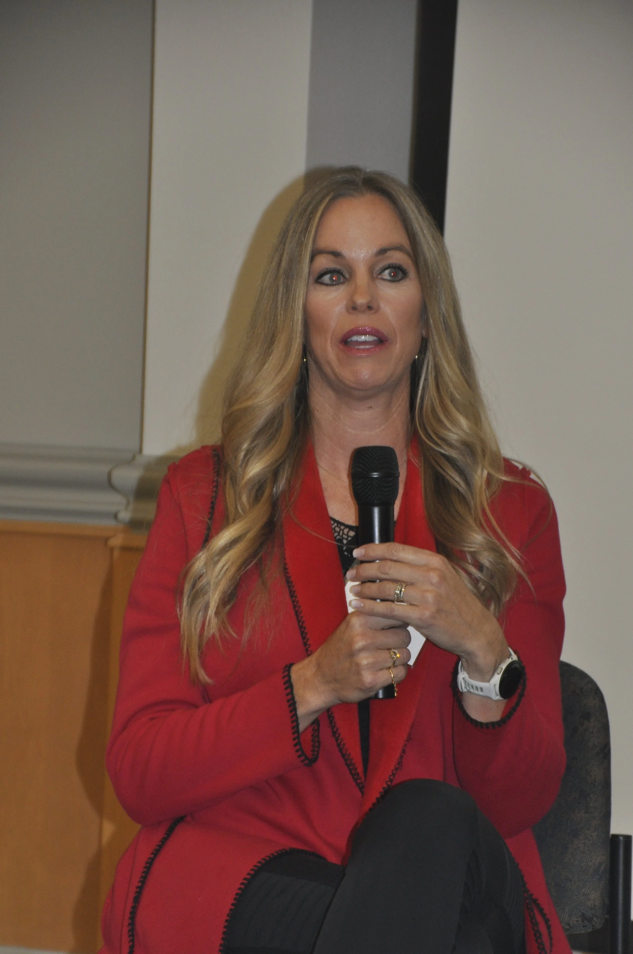 A woman with long blonde hair, wearing a red jacket, holding a microphone, sitting in a room with gray and beige walls.