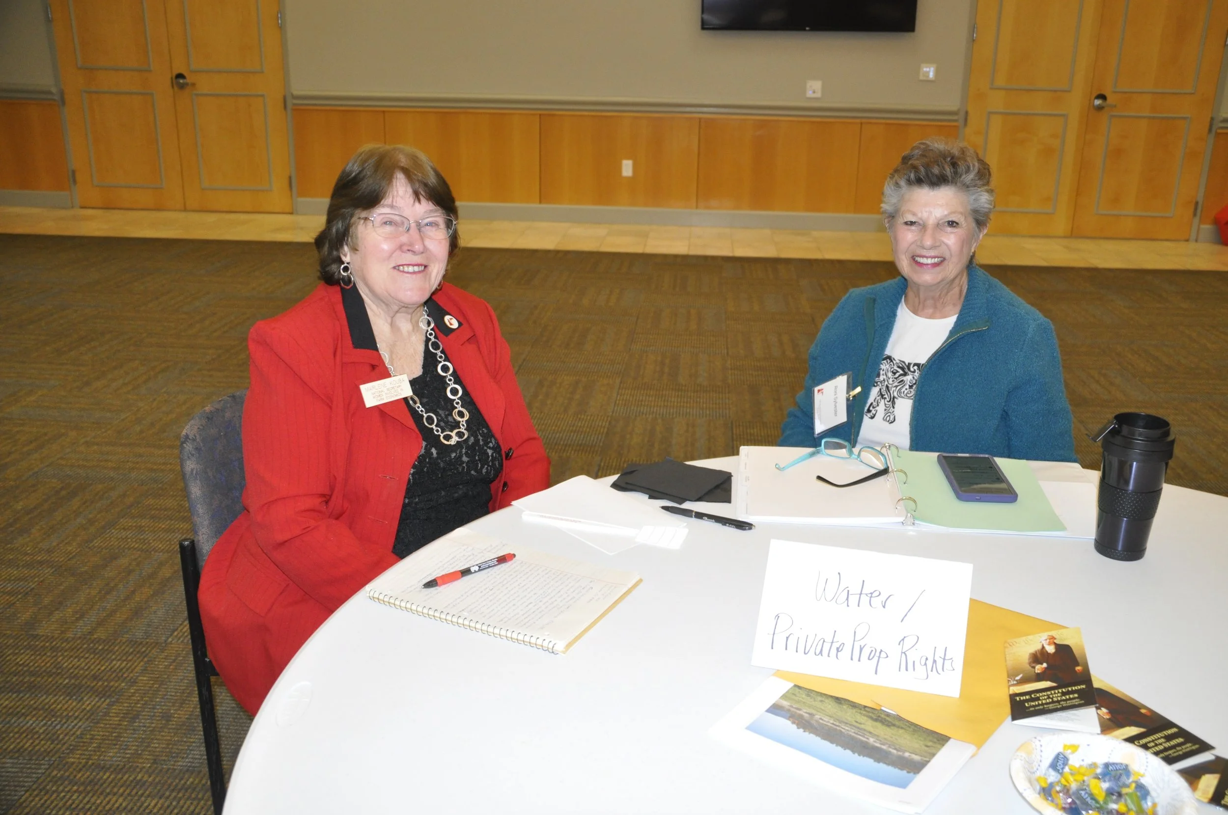 Two women sitting at a round table during an indoor event. The woman on the left is wearing a red jacket and glasses, and the woman on the right is wearing a teal jacket. The table has papers, a notebook, a pen, a smartphone, a cup, and a sign that r