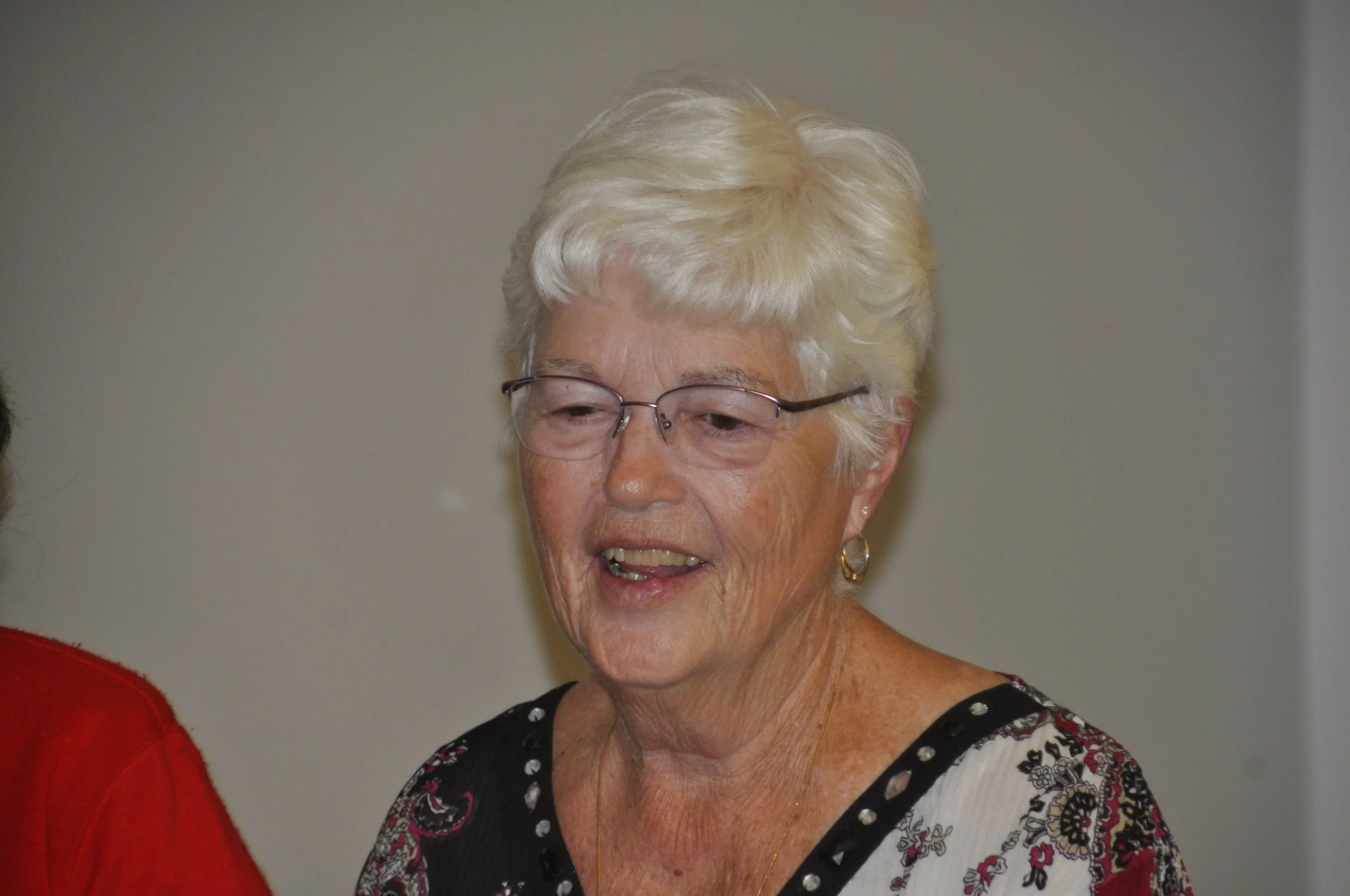 A close-up of an elderly woman with short, white hair, wearing glasses and gold hoop earrings, smiling while looking slightly downward. She is dressed in a black shirt with white, purple, and pink patterns.