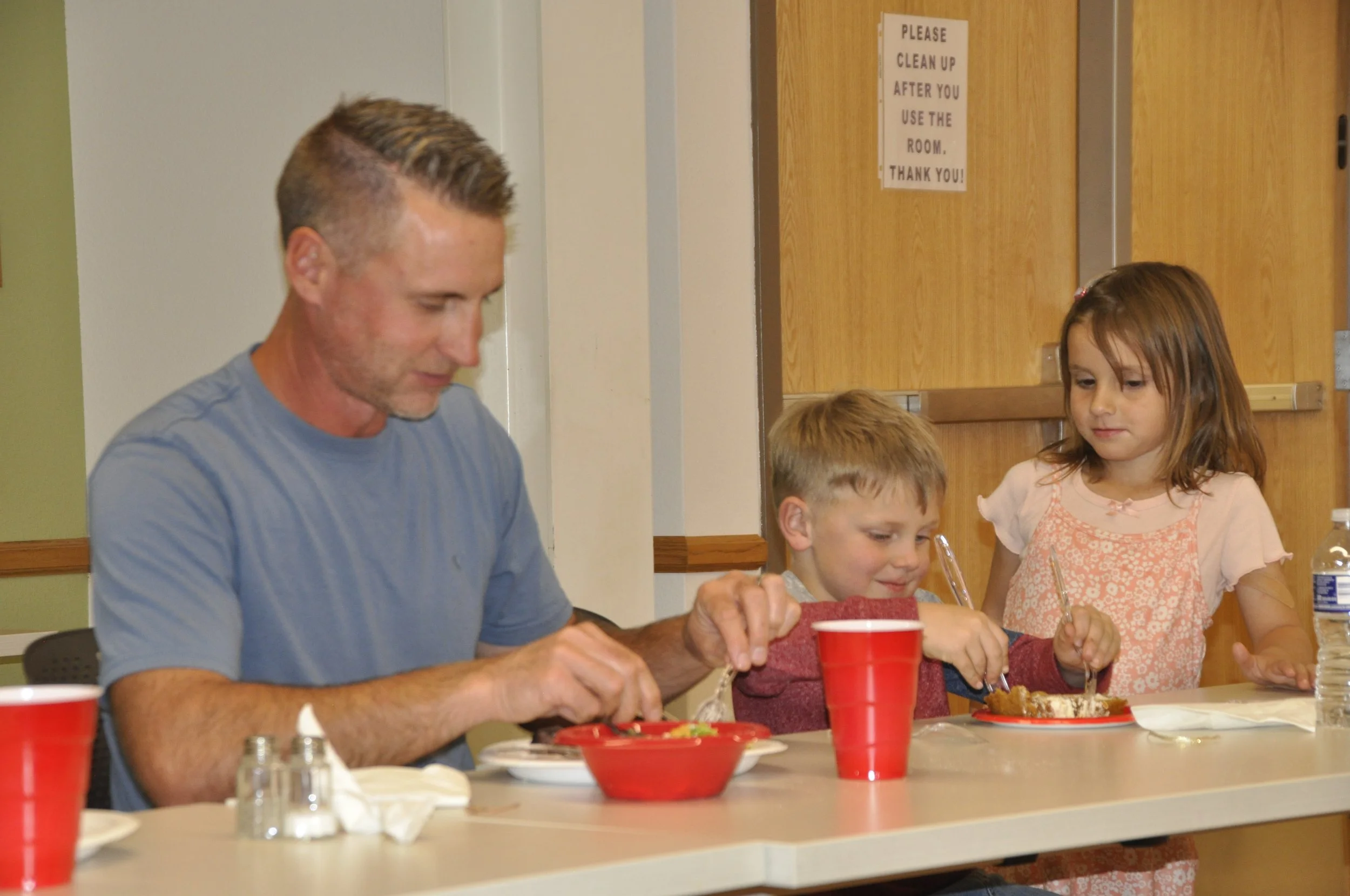 A group of people, including a man and two children, are sitting at a table eating food. There are red cups, bowls, and plates on the table. A sign on the door in the background reads, 'Please clean up after you use the room. Thank you!'