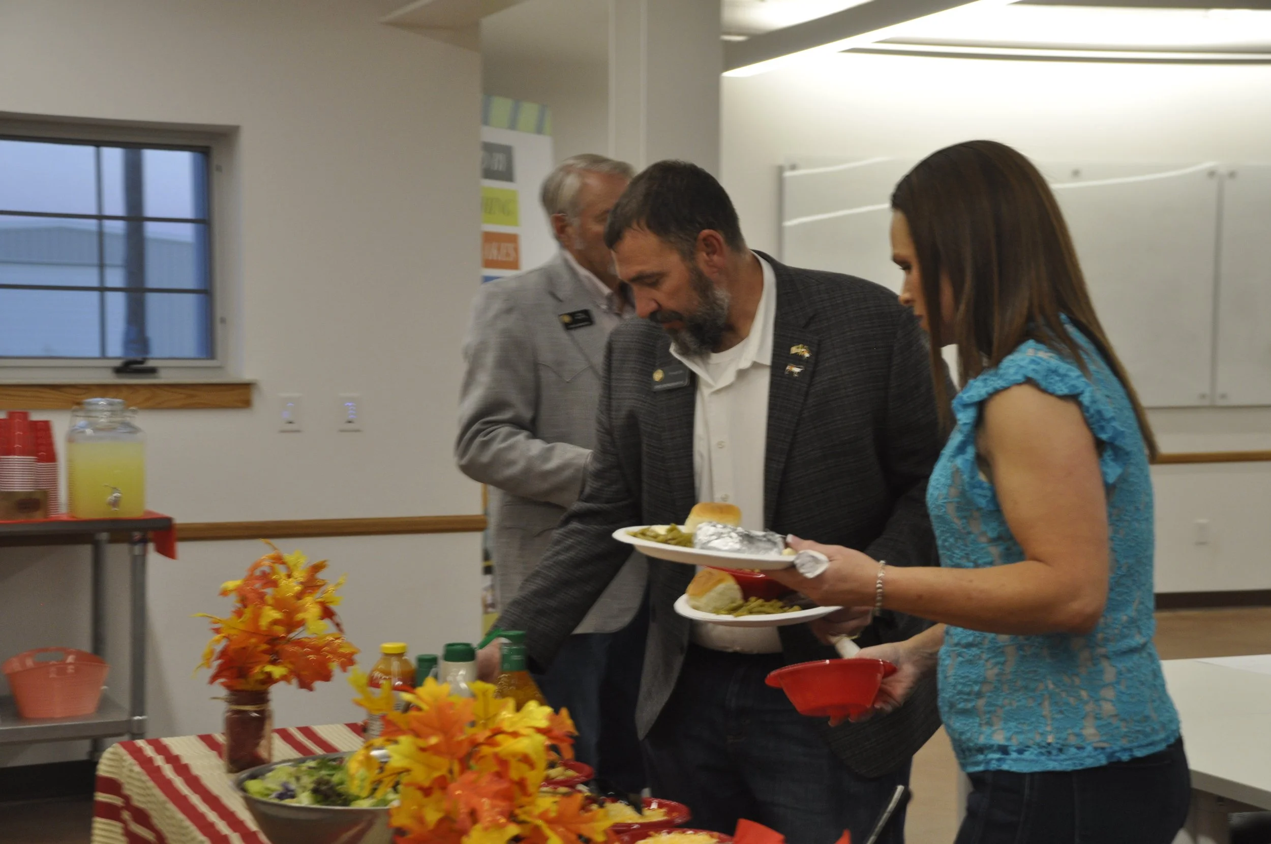 People attending a buffet in a decorated room with fall-themed decor, including a table with bowls of food, vegetables, and drinks, and colorful autumn leaves as centerpieces.