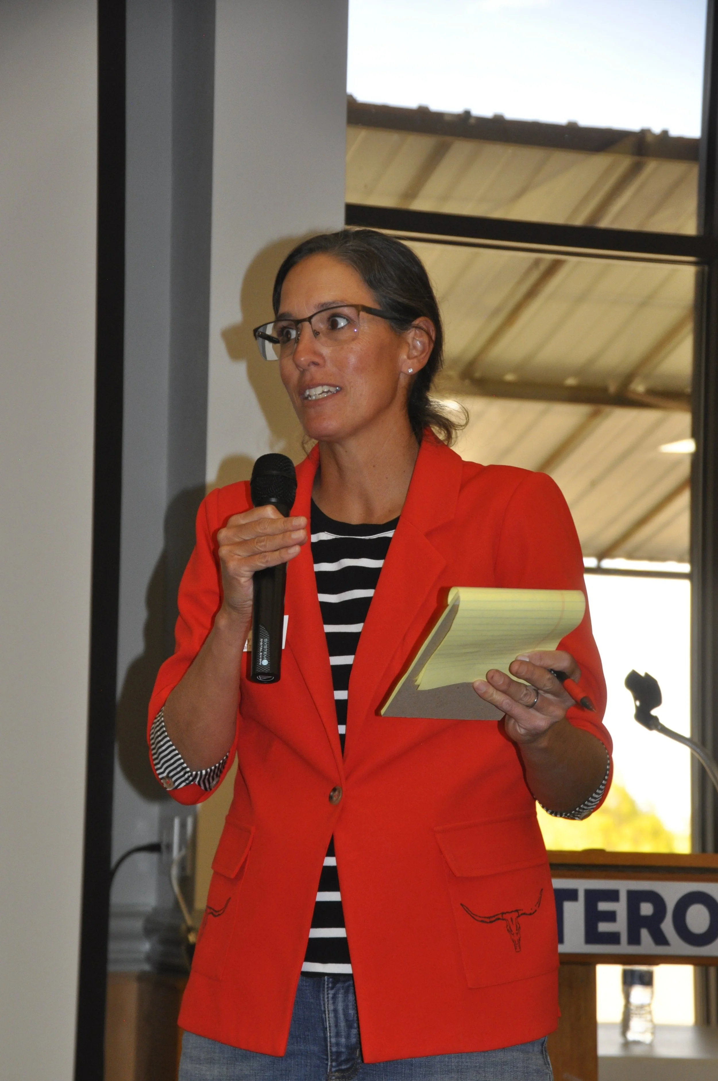 A woman wearing glasses and a red blazer with a black and white striped shirt underneath speaks into a microphone while holding a yellow notepad, standing in front of a window.
