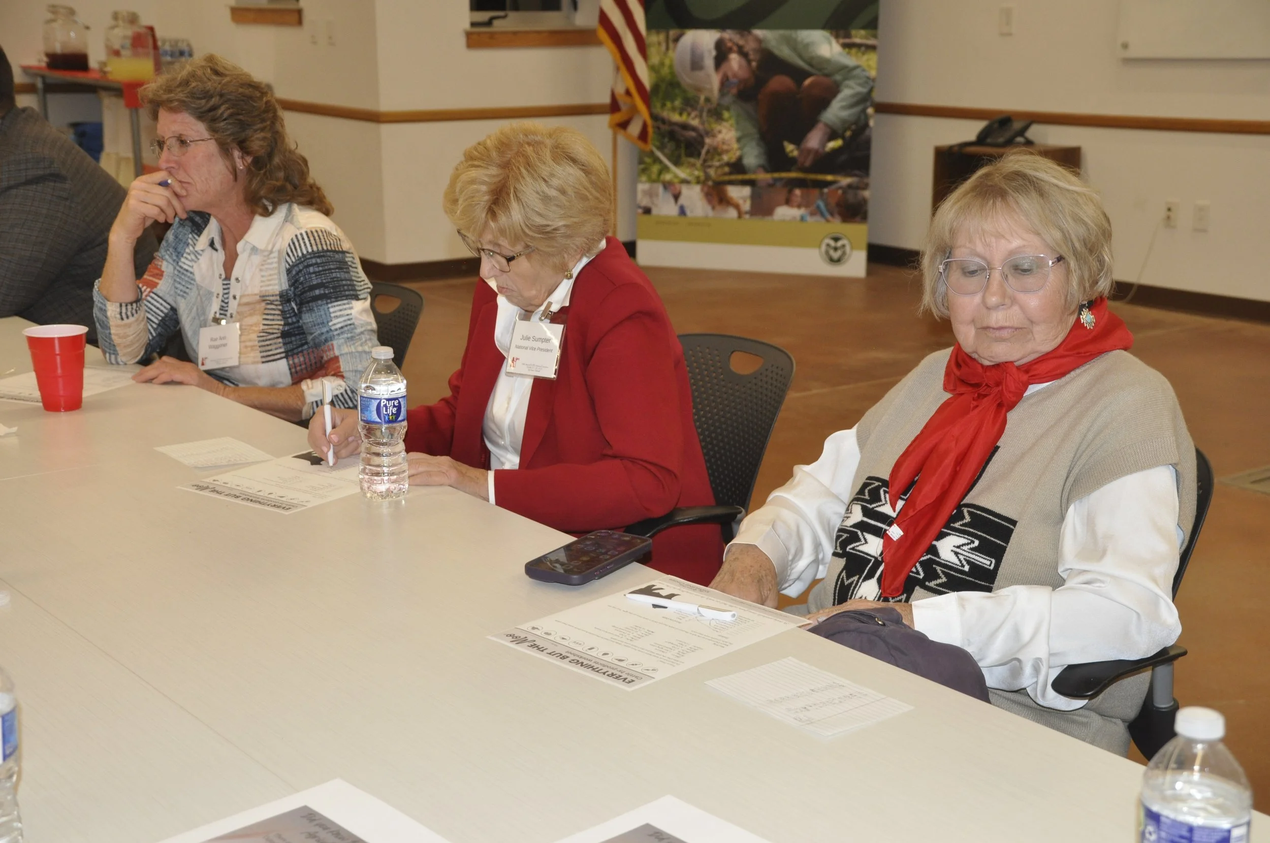 Three elderly women sitting at a long table during a meeting or conference, with papers and water bottles in front of them. They are engaged in reading or writing. There is a banner and American flag in the background.