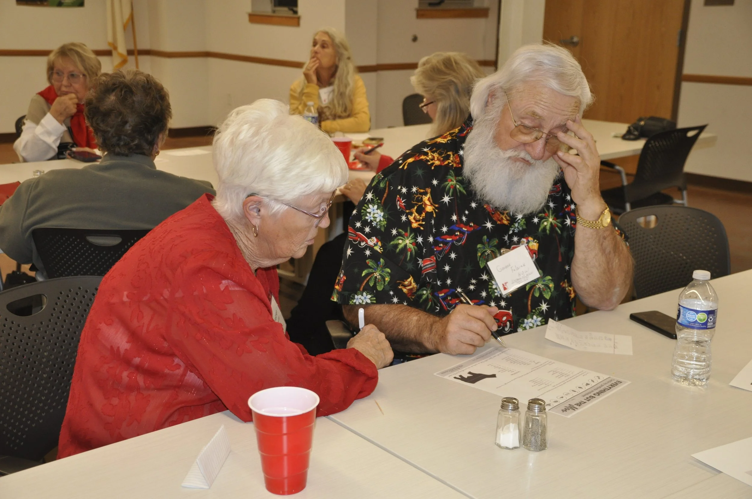 A group of elderly people sitting at a conference table, reading and writing on papers, with one man resting his head on his hand, wearing a festive, Christmas-themed shirt.