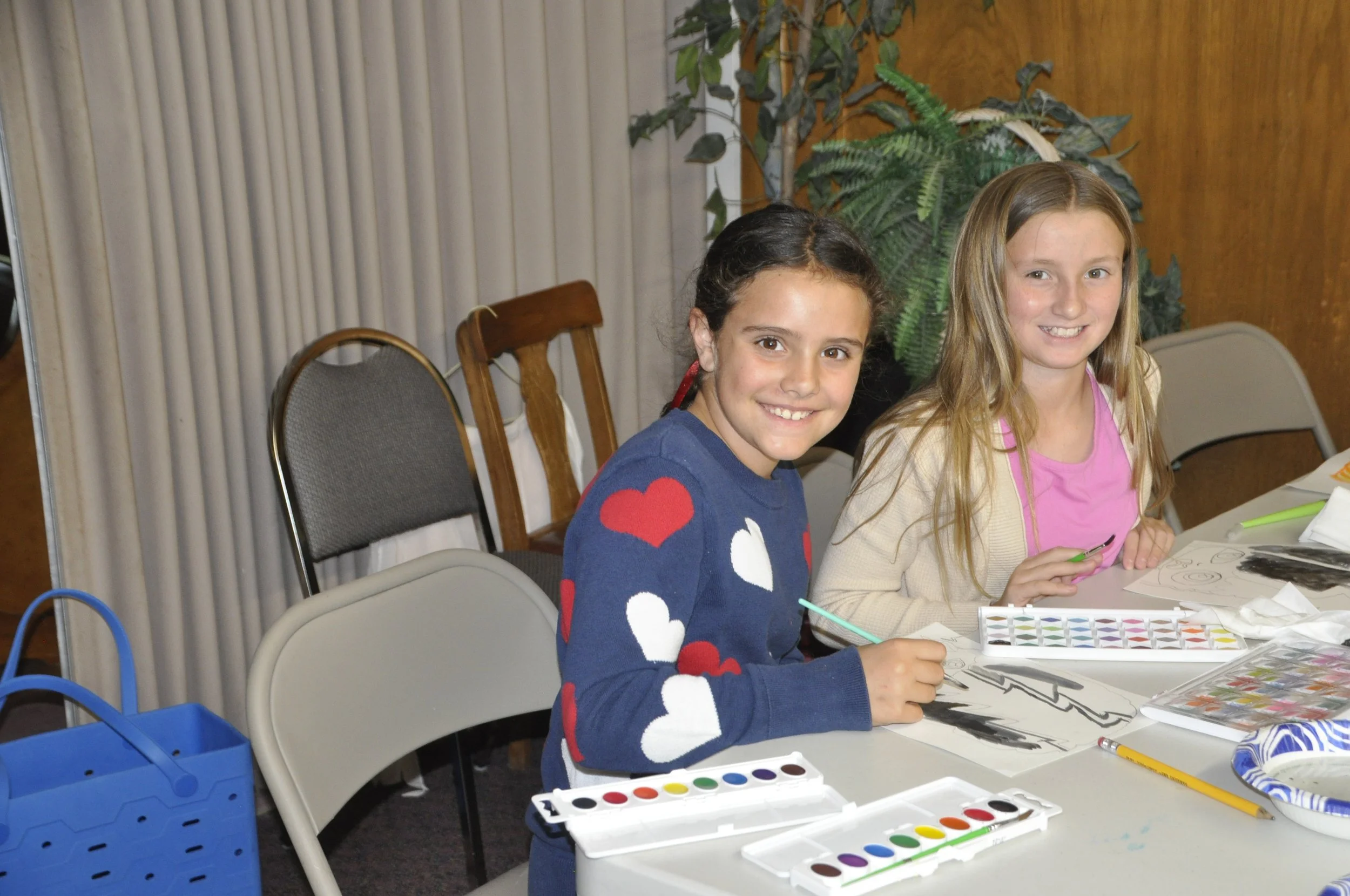 Two young girls sitting at a table, smiling at the camera, engaged in painting and drawing activities with watercolor paints, brushes, and papers in front of them.