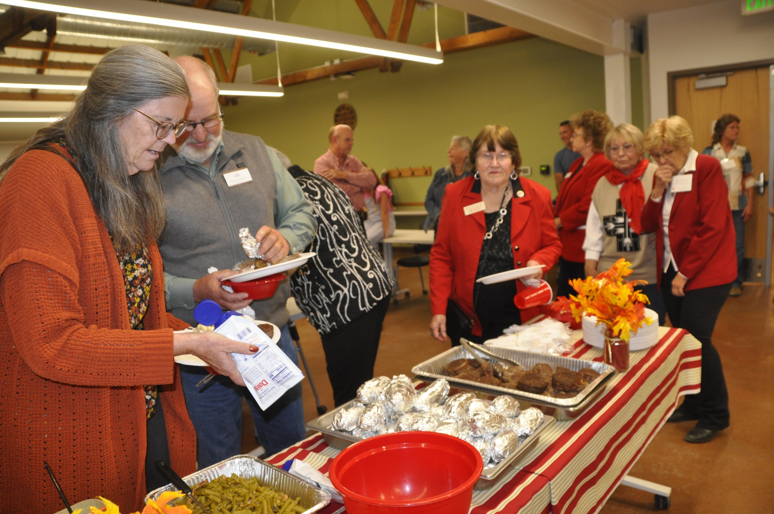 People gathered around a table with food at a social event. There are plates of baked goods, dishes, and a decorative centerpiece with autumn leaves.