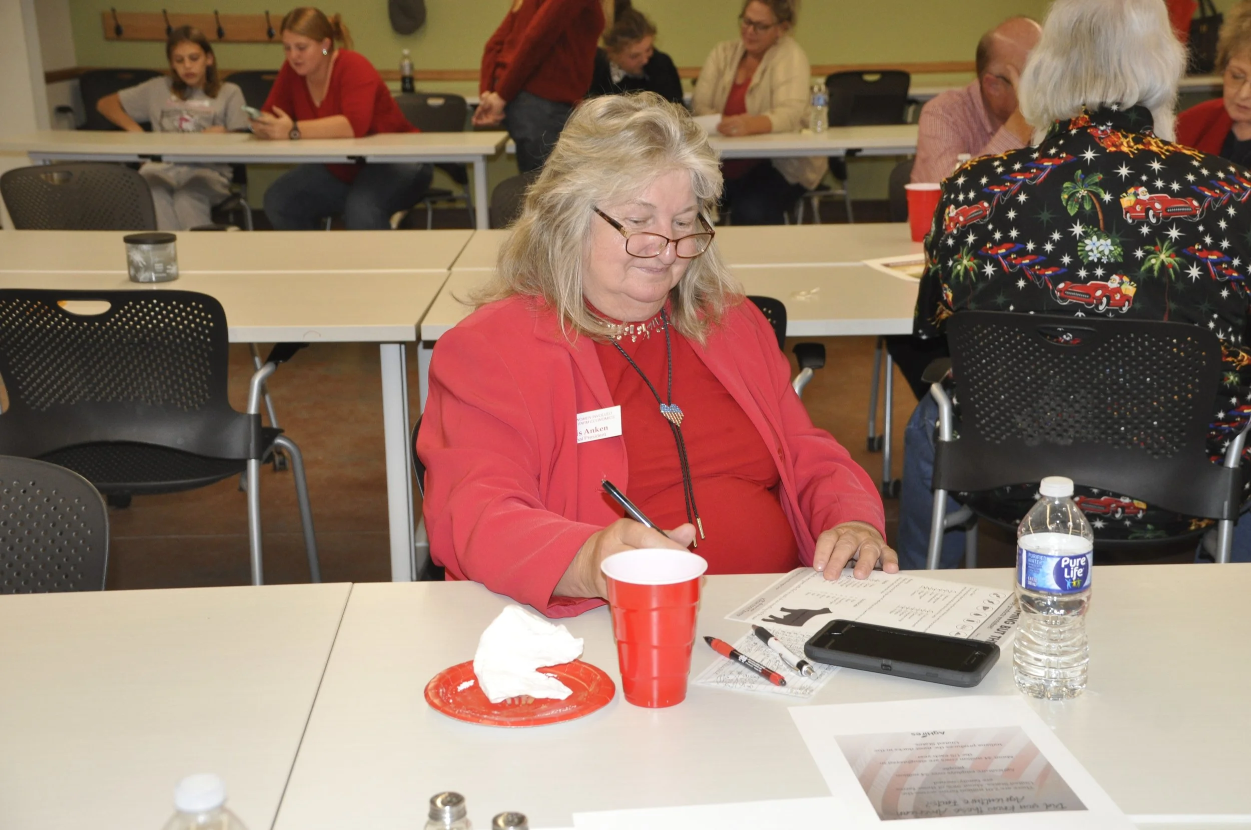 A group of people sitting at tables in a room, engaging in an activity. In the foreground, a woman with gray hair, glasses, and a red jacket is writing on a piece of paper, with a water bottle, a red cup, and some papers on the table. In the backgrou