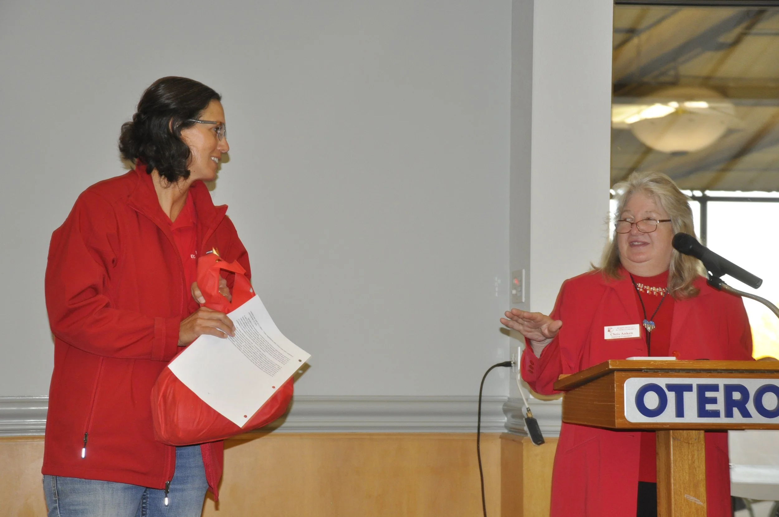 Two women at an event, both wearing red jackets, one holding a paper and the other near a podium with a microphone, engaged in conversation.
