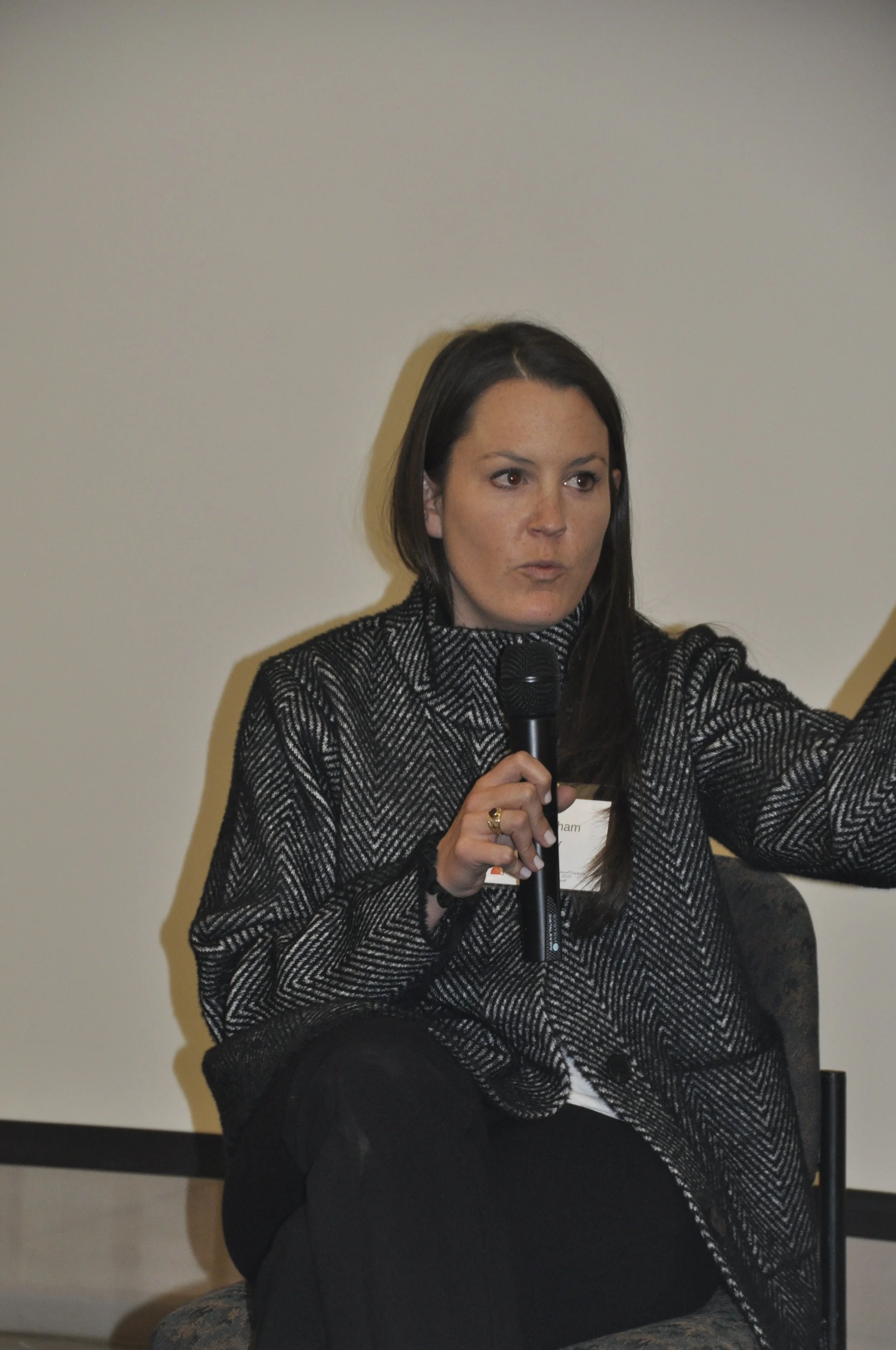 A woman with long dark hair and freckles, wearing a black and white patterned blazer, sitting down and holding a microphone while speaking at an indoor event.