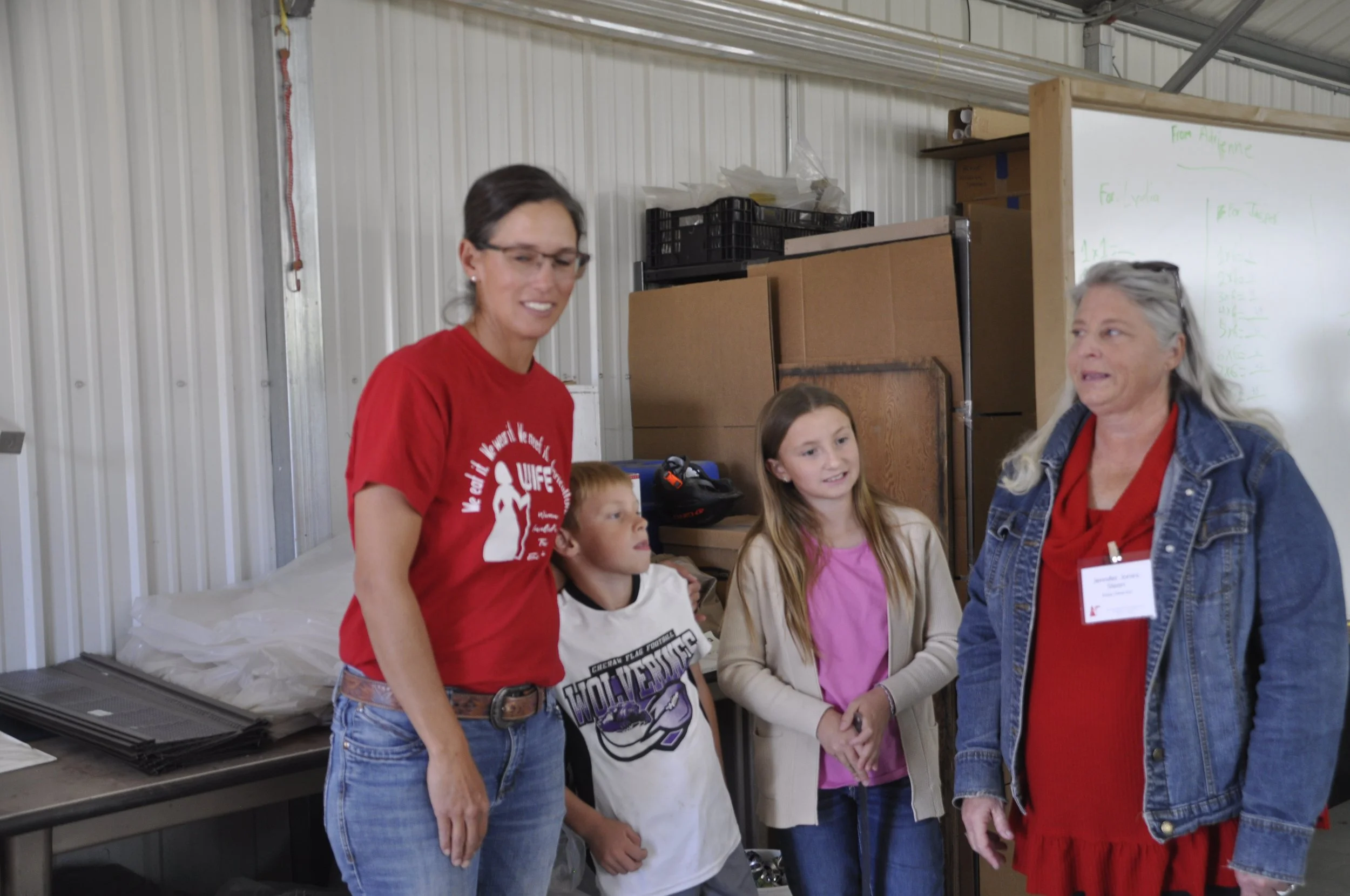 A group of five people, including children and adults, standing together in an industrial or warehouse setting, with a whiteboard and boxes in the background.
