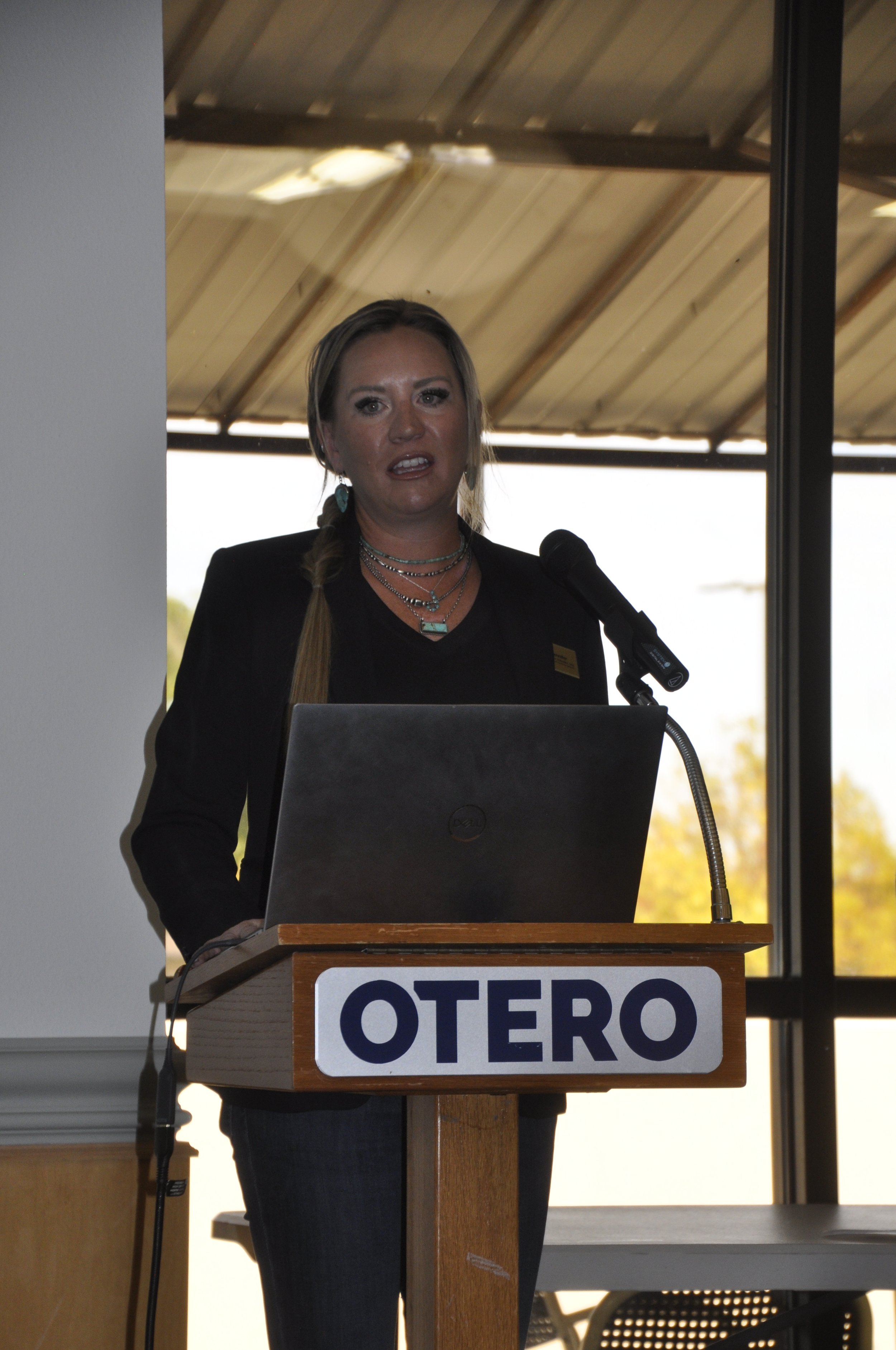 A woman with blonde hair and layered necklaces giving a speech at a podium with a sign reading "OTERO". She is indoors near large windows with a metal roof visible outside.
