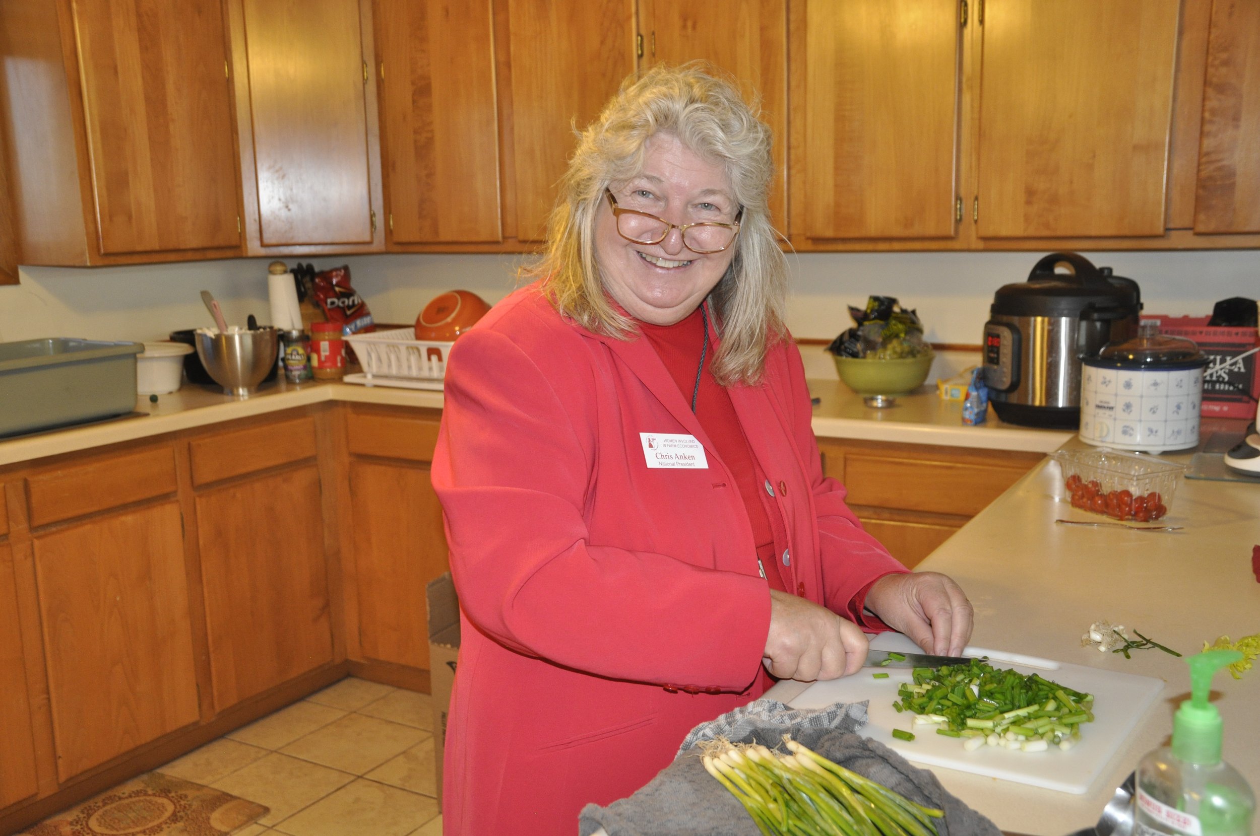 A woman with blonde hair wearing glasses and a red blazer is chopping green vegetables in a kitchen. There are wooden cabinets, a variety of kitchen items, and a bowl of cherry tomatoes on the counter.