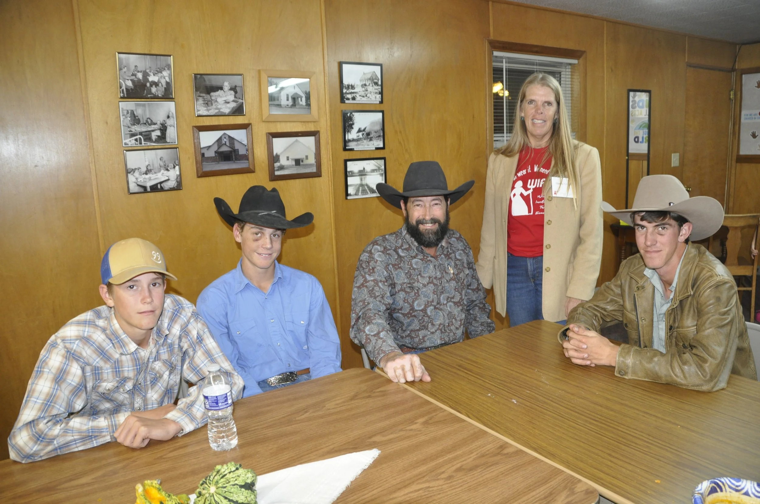 A group of five people, including three young men and two women, all wearing cowboy hats, sitting around a wooden table in a room with wood-paneled walls and framed pictures. The woman with long blonde hair and a tan blazer stands behind the group. T