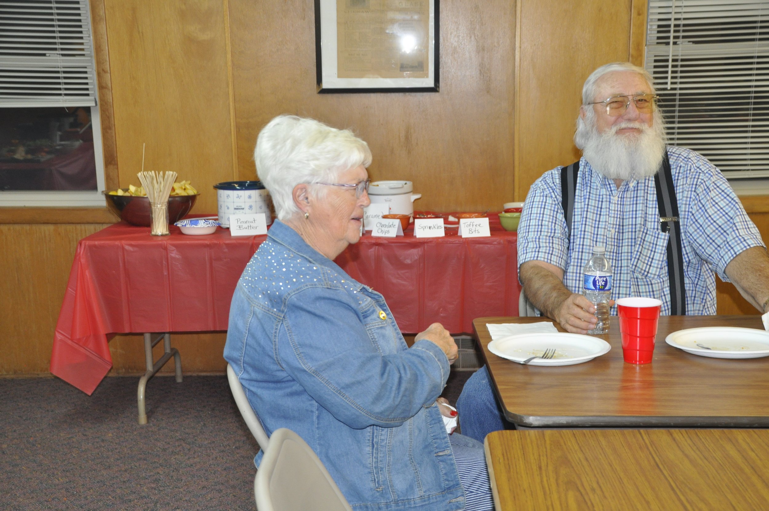 An elderly man and woman with white hair and beards sitting at a table during a social gathering. The man is smiling and holding a water bottle, while the woman is looking down and appears to be talking or listening. In the background, there is a tab