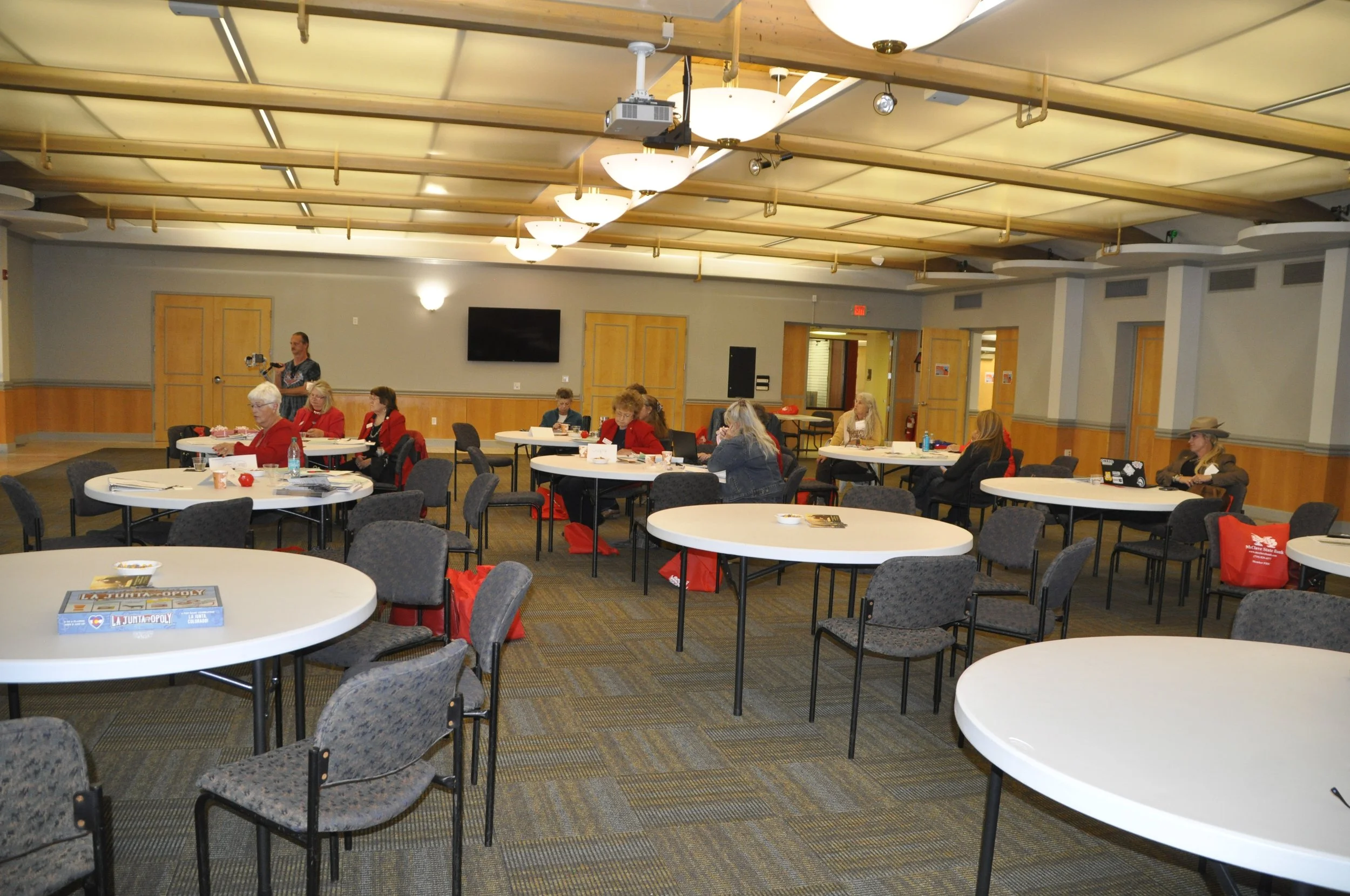 A conference room with round tables and gray chairs, some women seated at the tables, a woman standing and recording with a camera, wooden doors, a black TV screen, and ceiling lights.