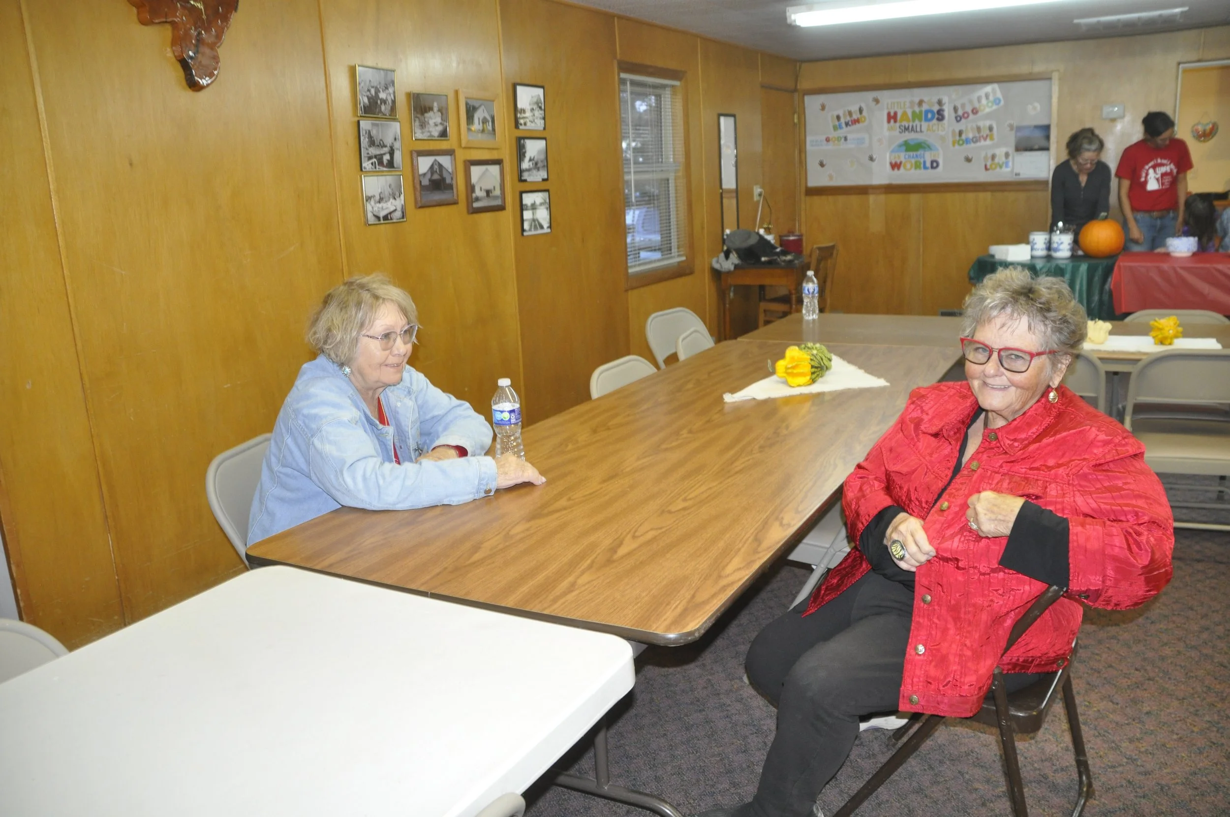 Two elderly women sitting at a long wood table in a room with wood-paneled walls, one in a light blue jacket and glasses, the other in a red jacket and glasses, smiling. In the background, a group of people is gathered around a table with a pumpkin o