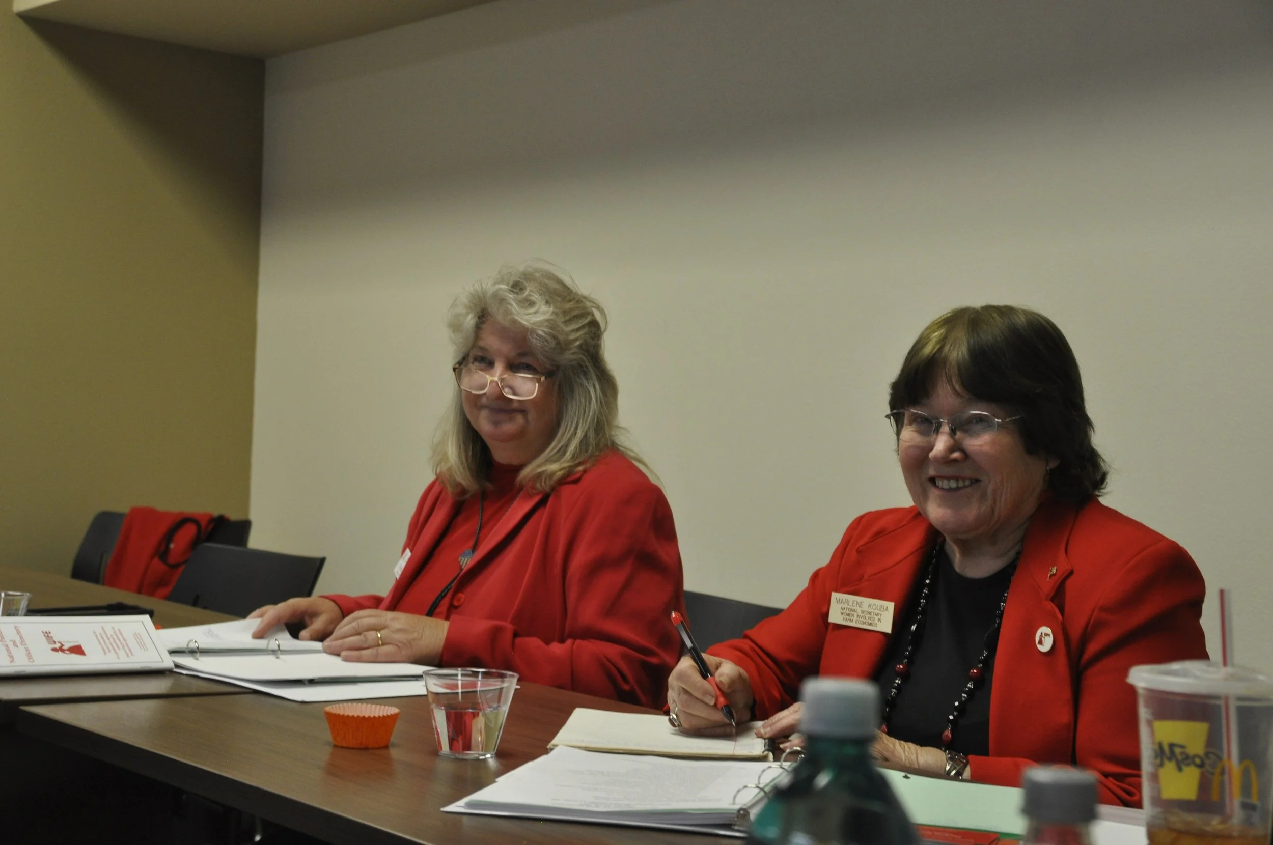 Two women in red blazers sitting at a conference table, smiling, with papers, pens, and beverages in front of them. The woman on the left has blonde hair and glasses, while the woman on the right has dark hair and glasses and is taking notes.