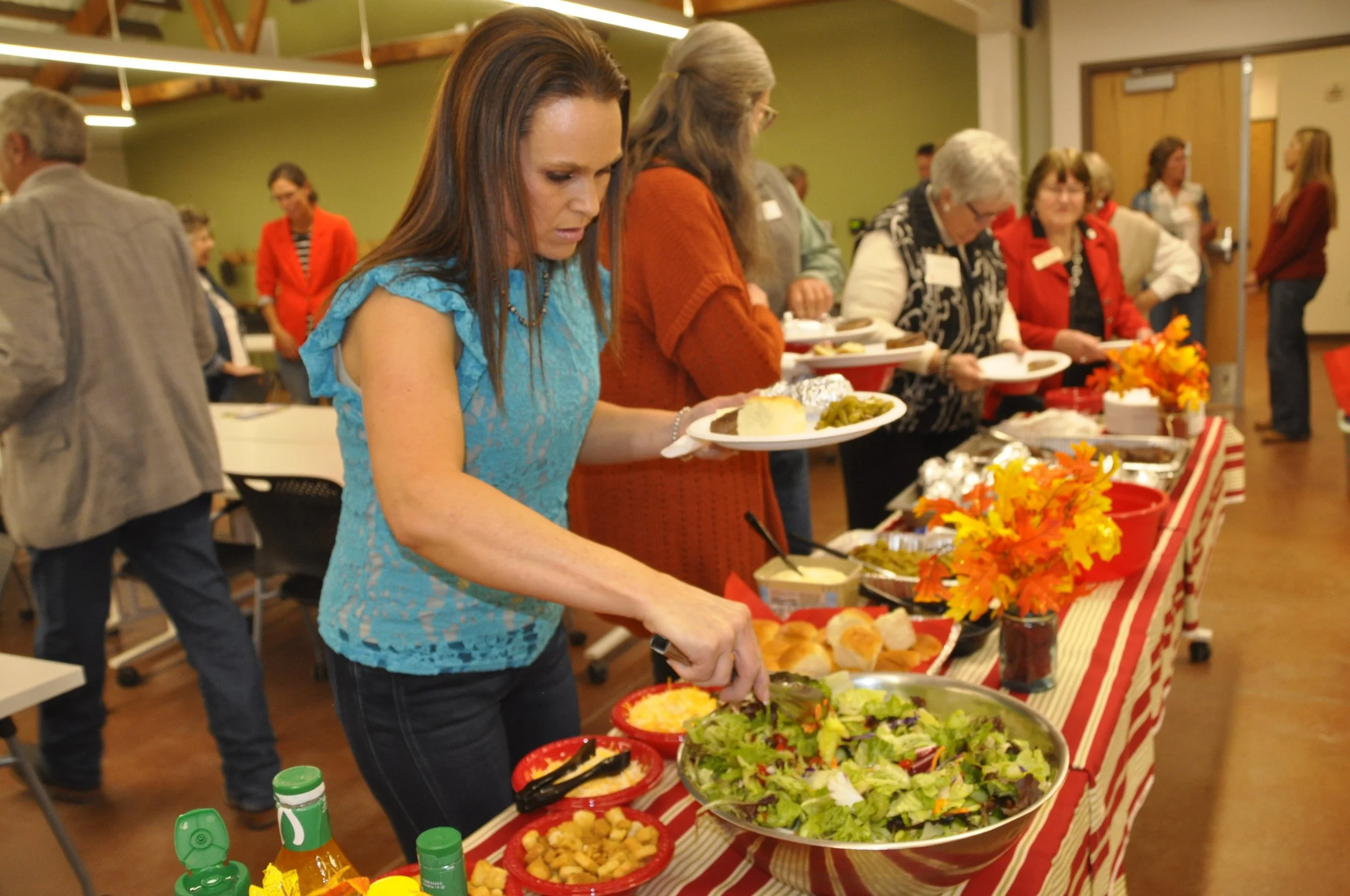 People standing in line at a buffet table filled with salad, bread, and various dishes, in a room decorated with fall-themed decorations and orange flowers.